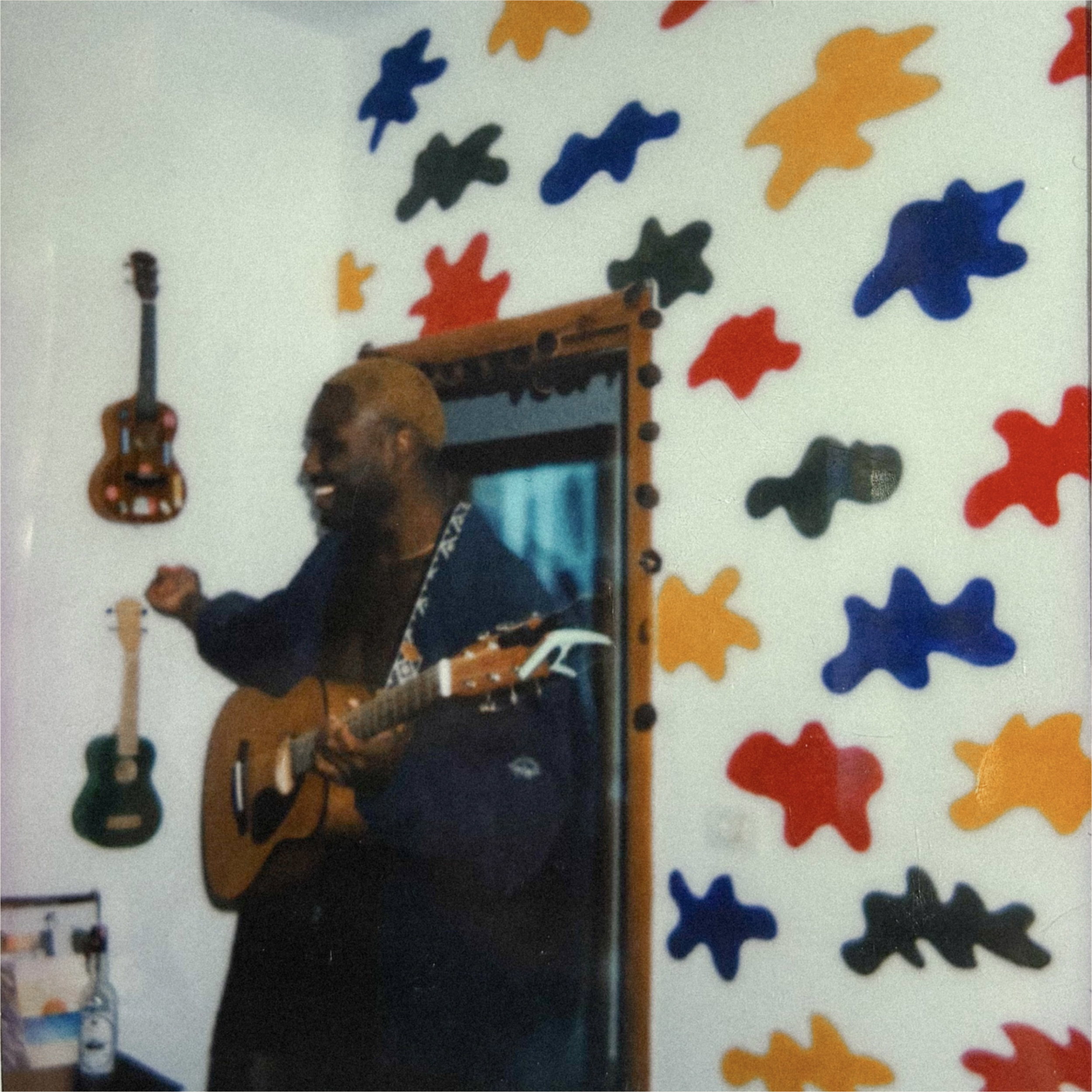A man with blond hair playing an acoustic guitar indoors, smiling, surrounded by colorful abstract wall decorations resembling puzzle pieces in red, blue, yellow, and black, with small guitars hanging on the wall.