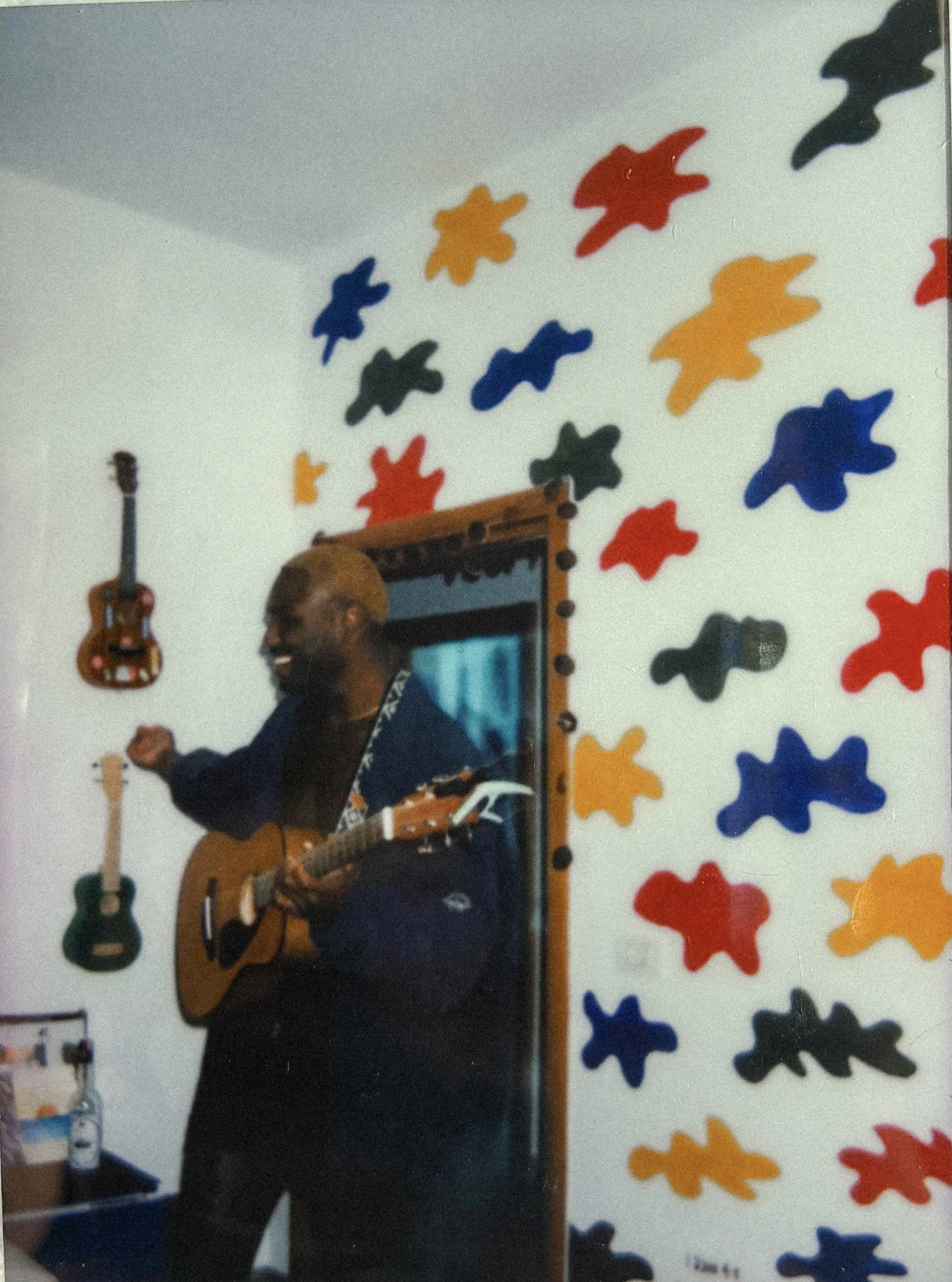 A person playing guitar in a room decorated with colorful abstract shapes on the wall.