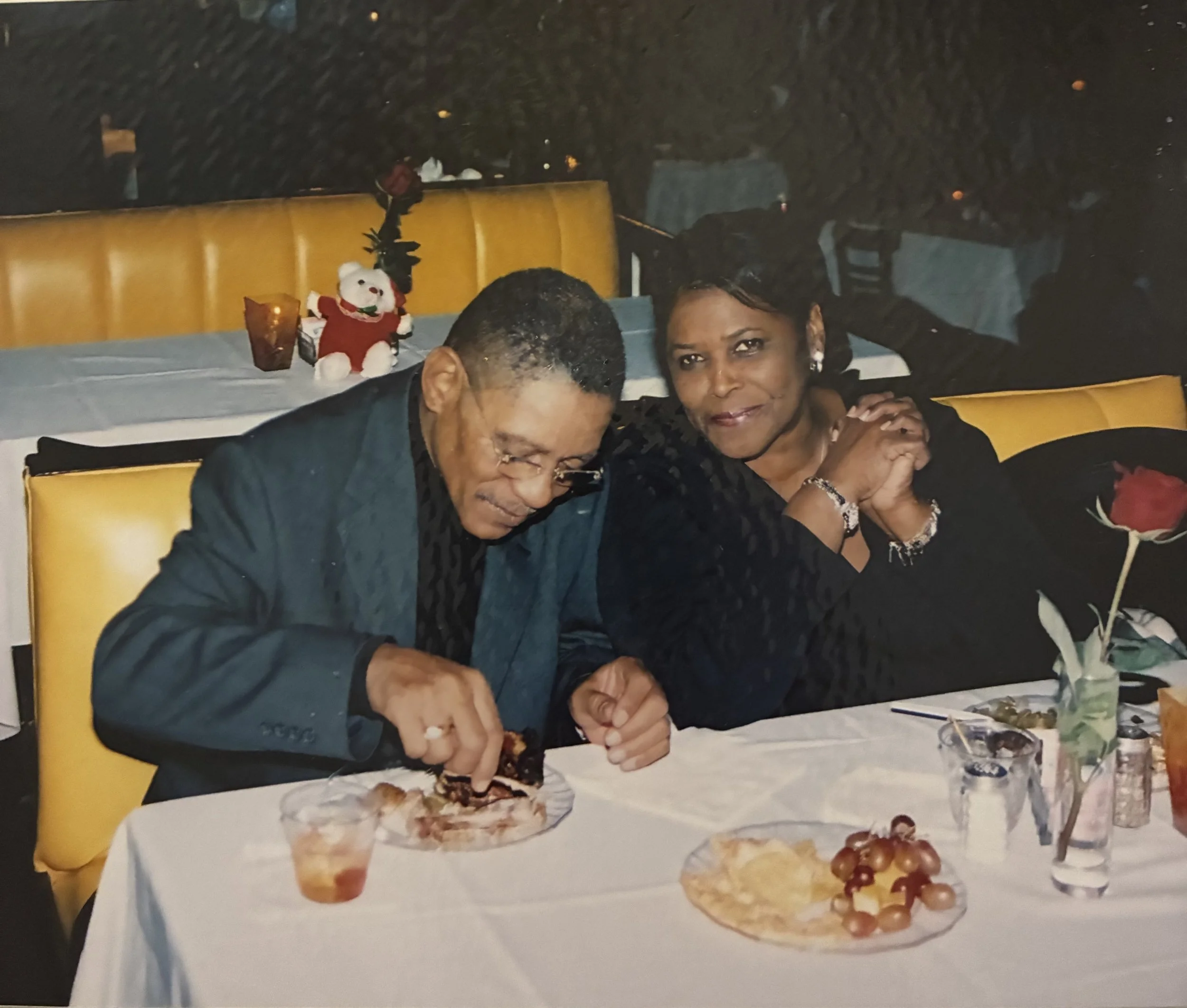 A man and woman sitting at a decorated dinner table with food, drinks, and a single red rose in a vase, in a restaurant setting with dark background.