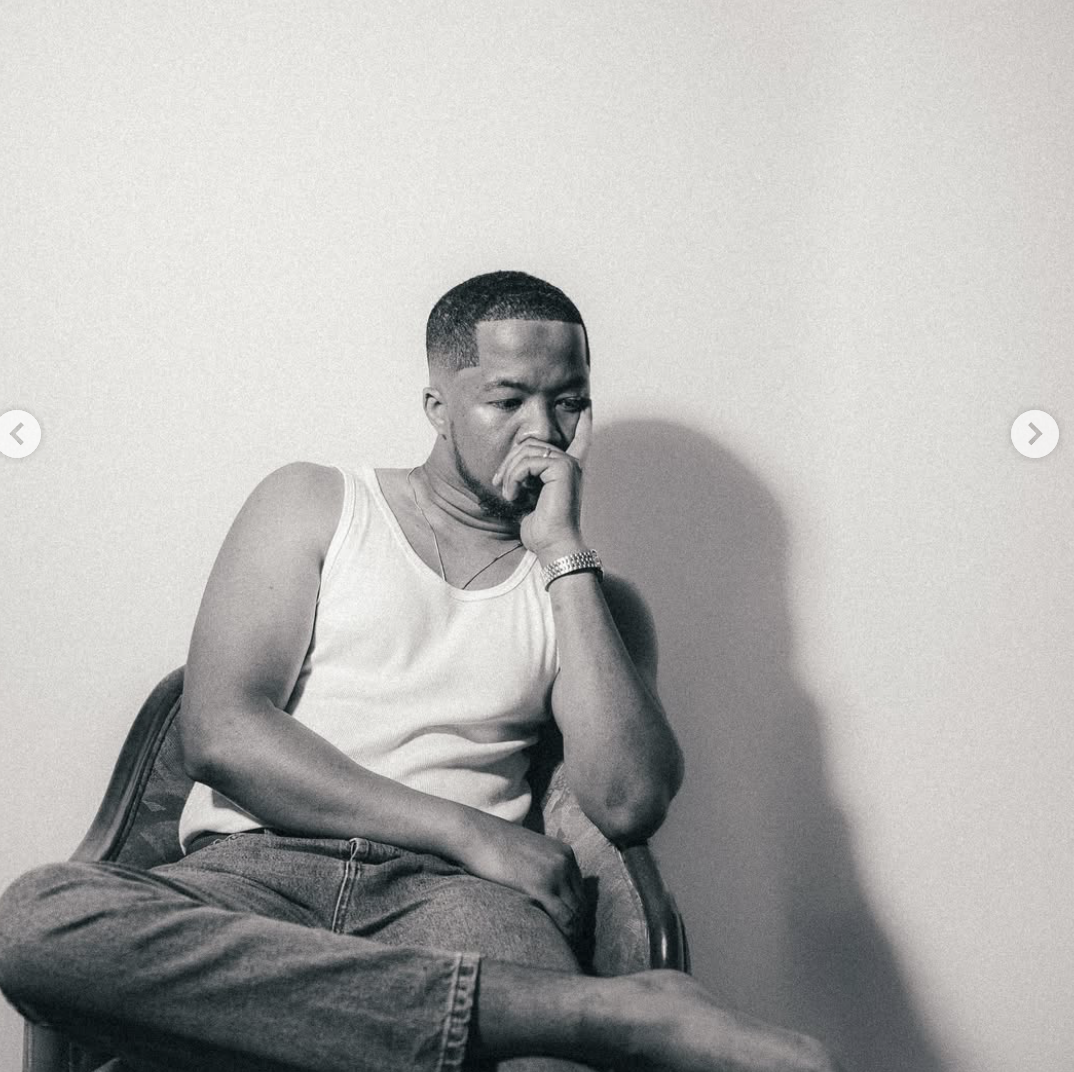 A young man with a short haircut sitting on a chair, wearing a white tank top, jeans, and a watch, with his hand resting on his chin, appearing thoughtful against a plain background.
