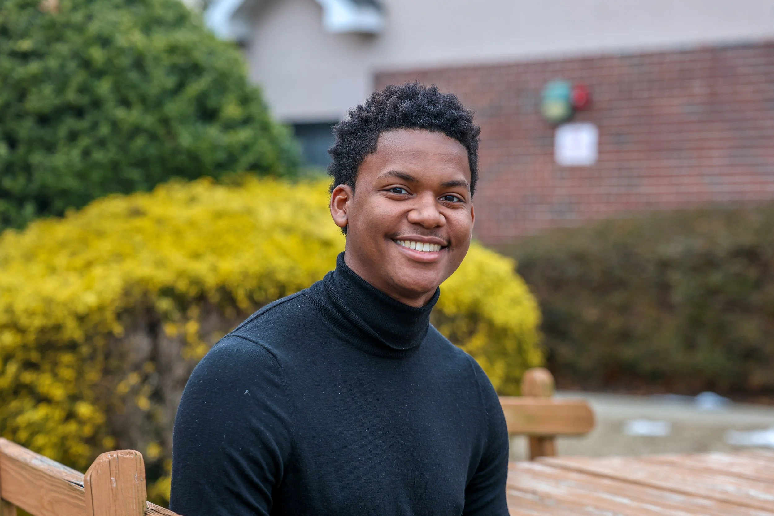A young man smiling outdoors, wearing a black turtleneck sweater, with a background of green and yellow shrubs and a brick building.