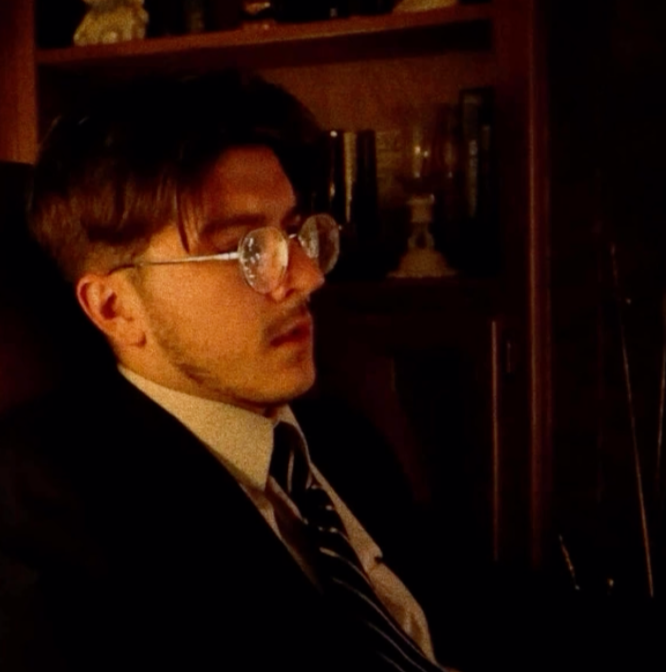 A man with glasses and brown hair, wearing a suit and tie, sitting indoors with a wooden bookshelf in the background.