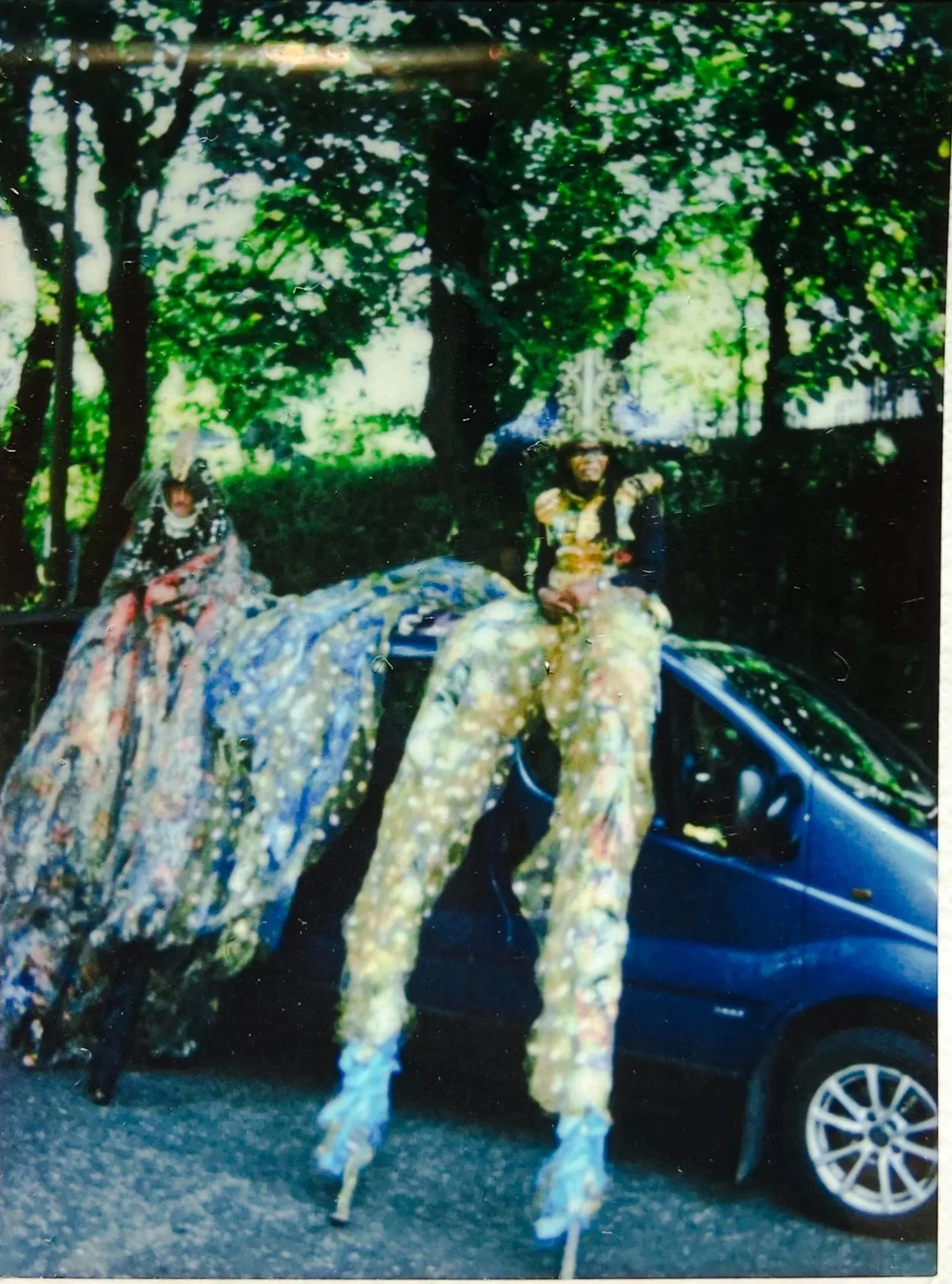 Two people dressed in elaborate colorful costumes with feathers and beads, sitting on the side of a black car parked on a street with green leafy trees in the background.