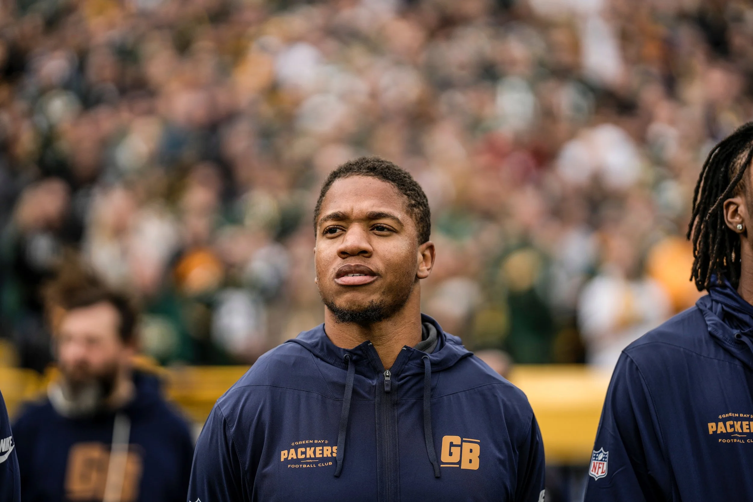A male football player wearing a navy blue Green Bay Packers jacket standing outdoors with a blurred crowd in the background during a game or event.