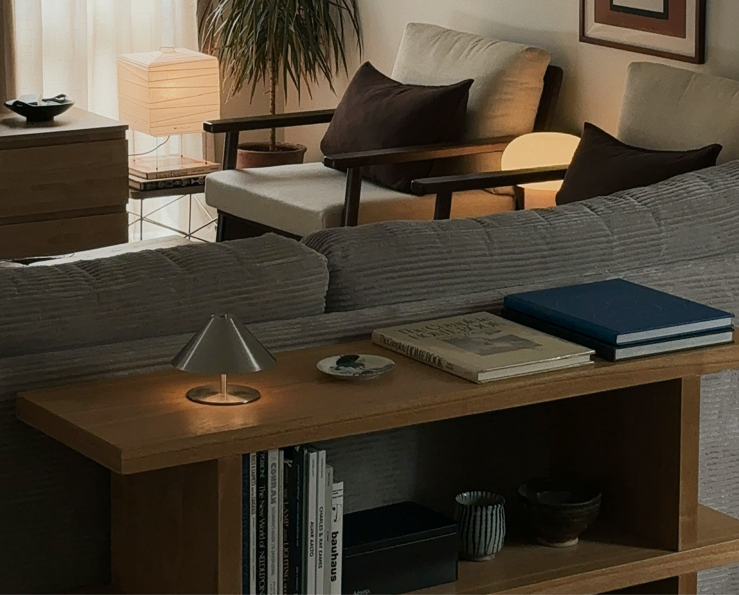 Living room with beige couch, wooden side table, and armchairs with dark cushions in front of a window with curtains. A small lamp on the side table and a table lamp behind the couch provide warm lighting. Books are on the coffee table and shelves.