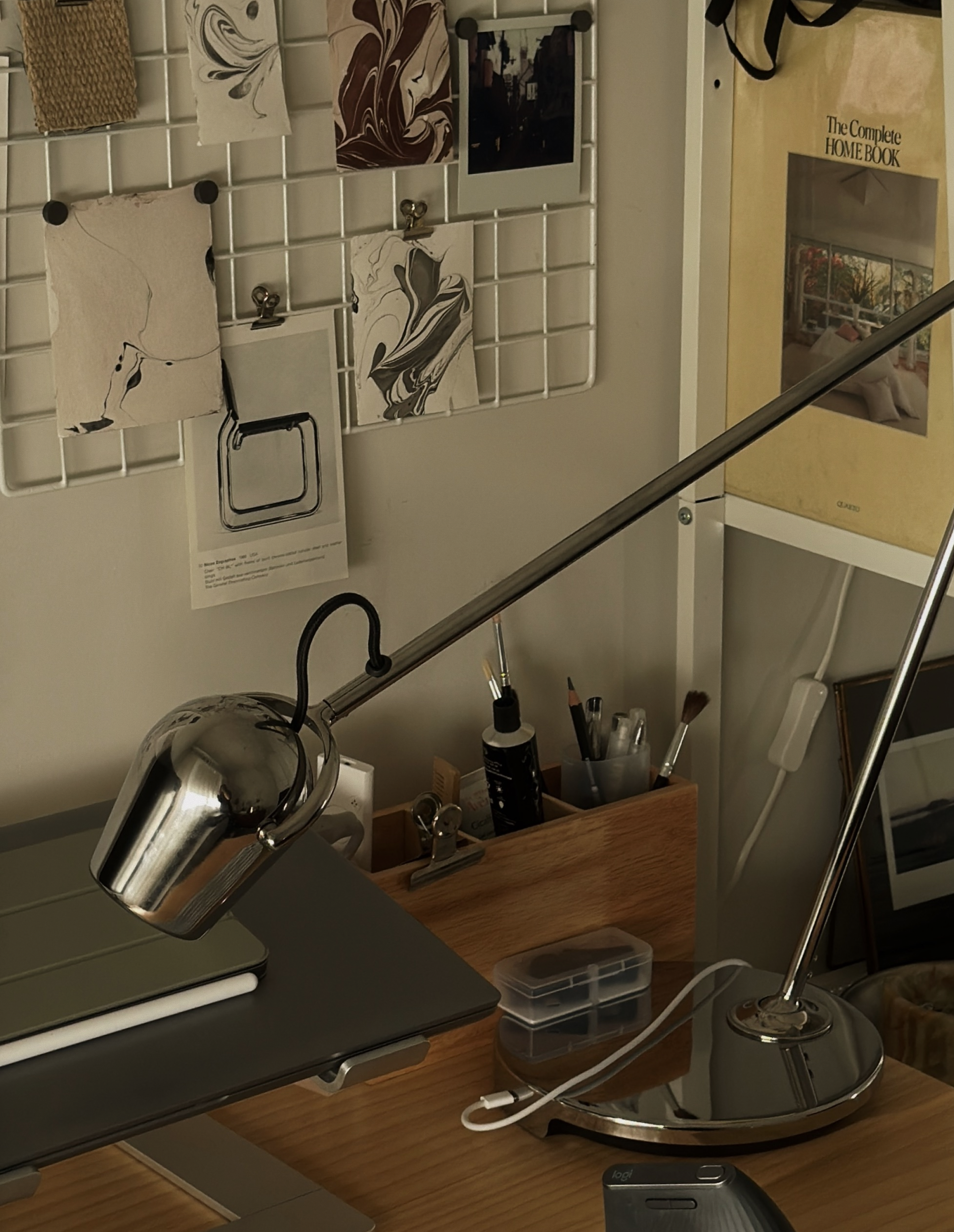 A desk with a silver adjustable desk lamp, a closed laptop, and various stationery items in a wooden organizer. In the background, there is a wall-mounted white grid displaying small artworks and photographs, and a bookshelf with a book titled 'The Complete Home Book.'