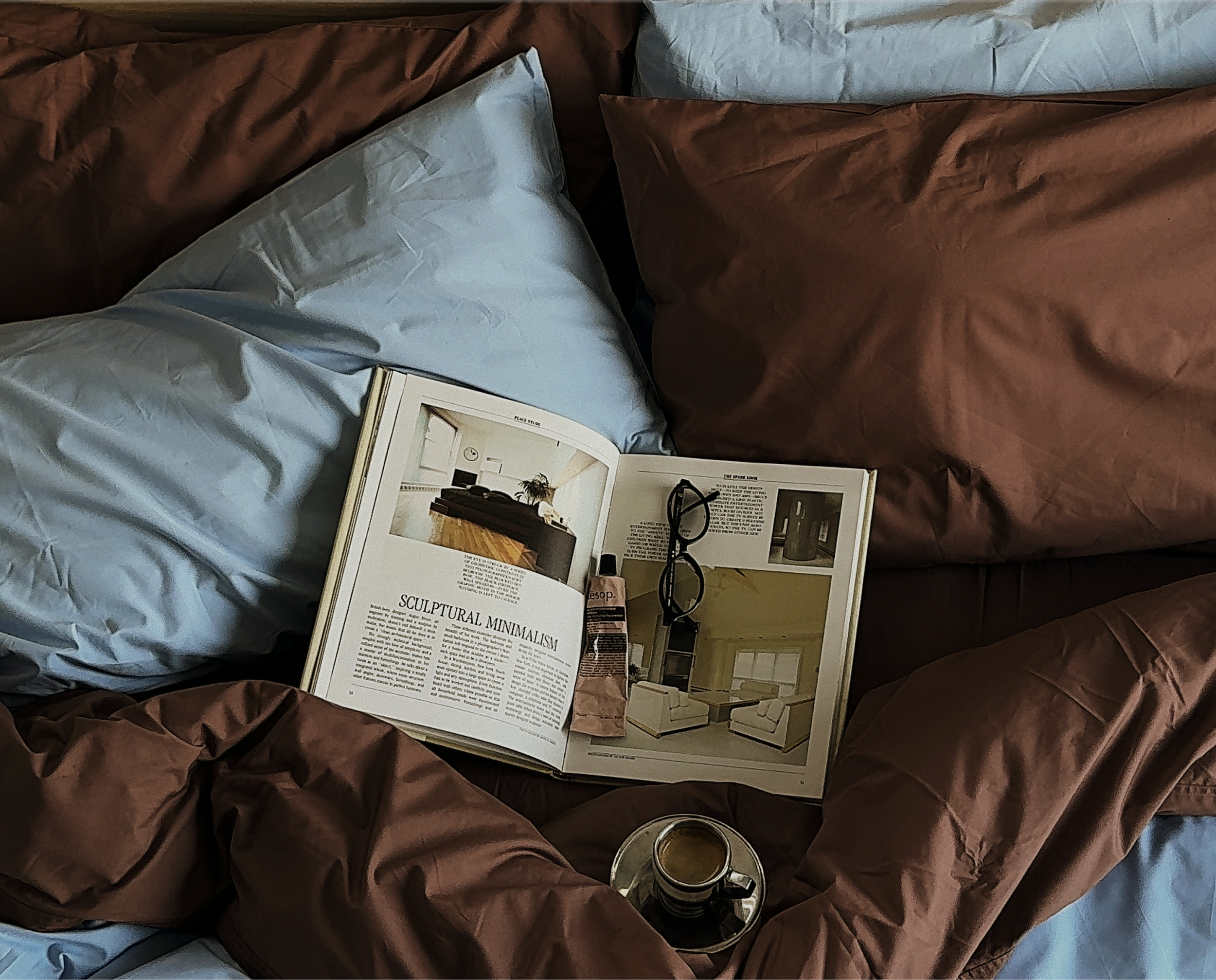 An open magazine resting on a bed with light blue and brown sheets, a pair of black glasses perched on the magazine, and a mug placed on the bed.