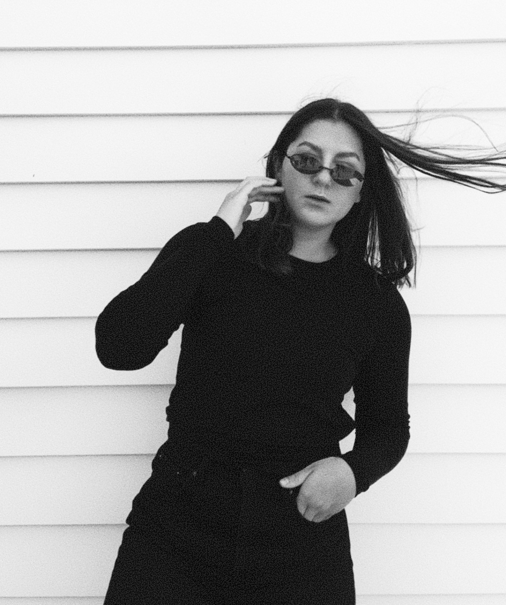 Black and white photo of a woman with long dark hair wearing sunglasses and a black outfit, standing against a white horizontal panel background, with wind blowing her hair. Mojan Mosavat