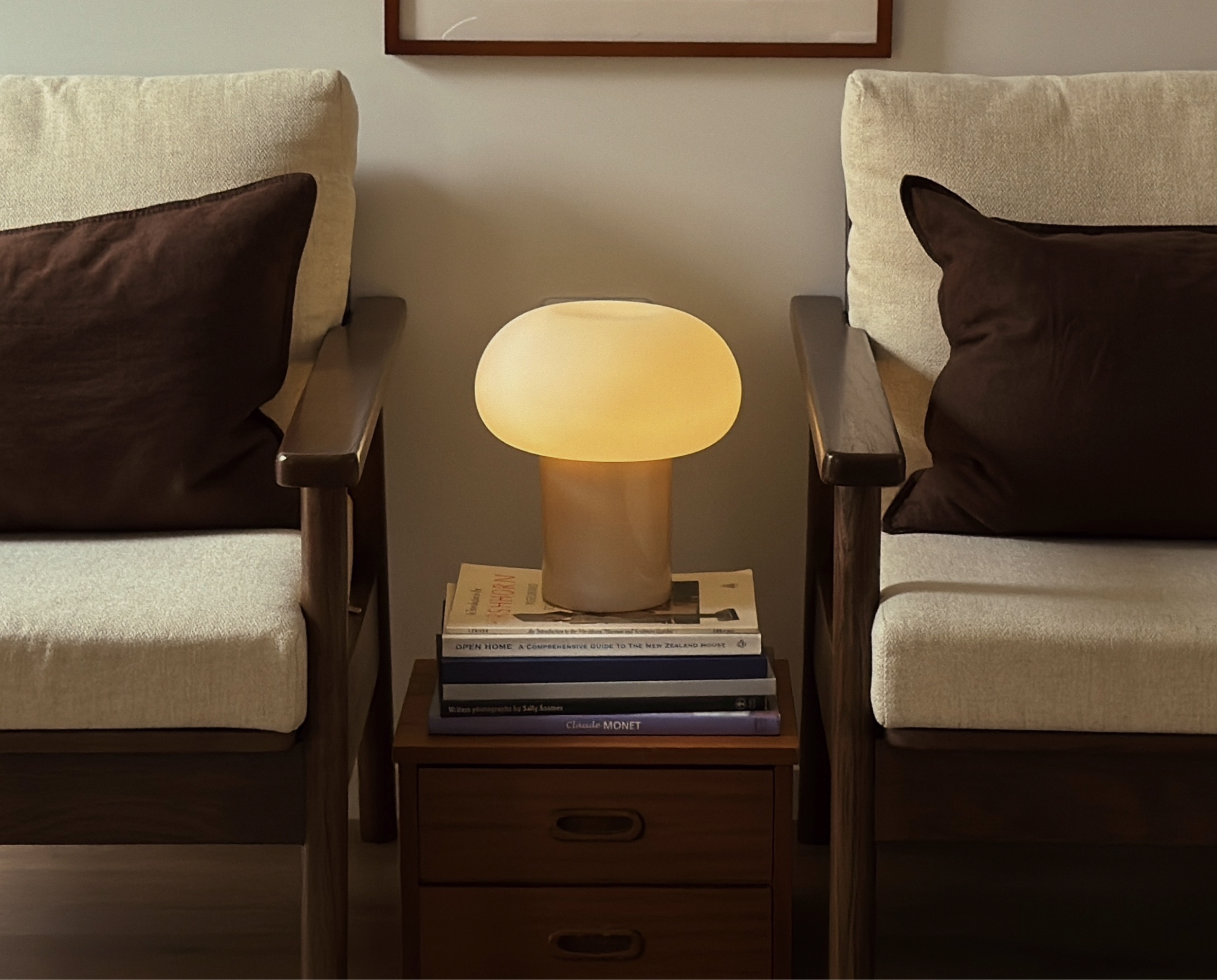 A bedroom with two beige armchairs with dark brown pillows, a wooden nightstand with three drawers, a luminous lamp shaped like a mushroom on top of the nightstand, and a framed picture hanging on the wall behind the chairs.