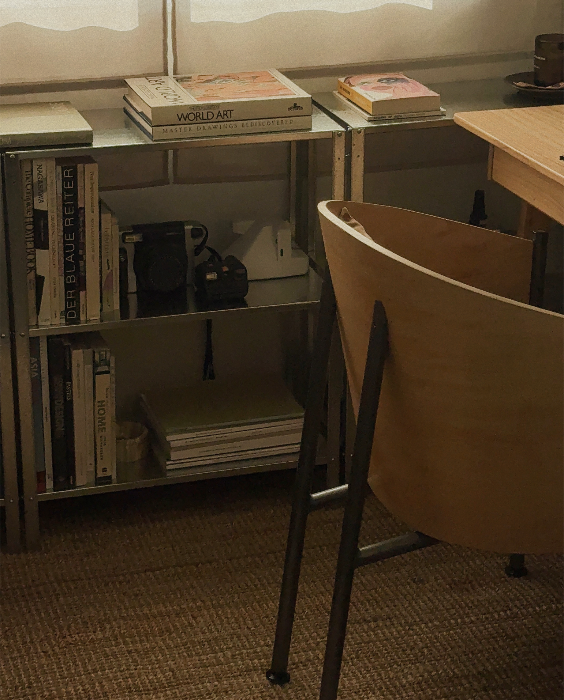 A metal bookshelf with books, magazines, and a camera, next to a wooden desk and chair in a cozy room.