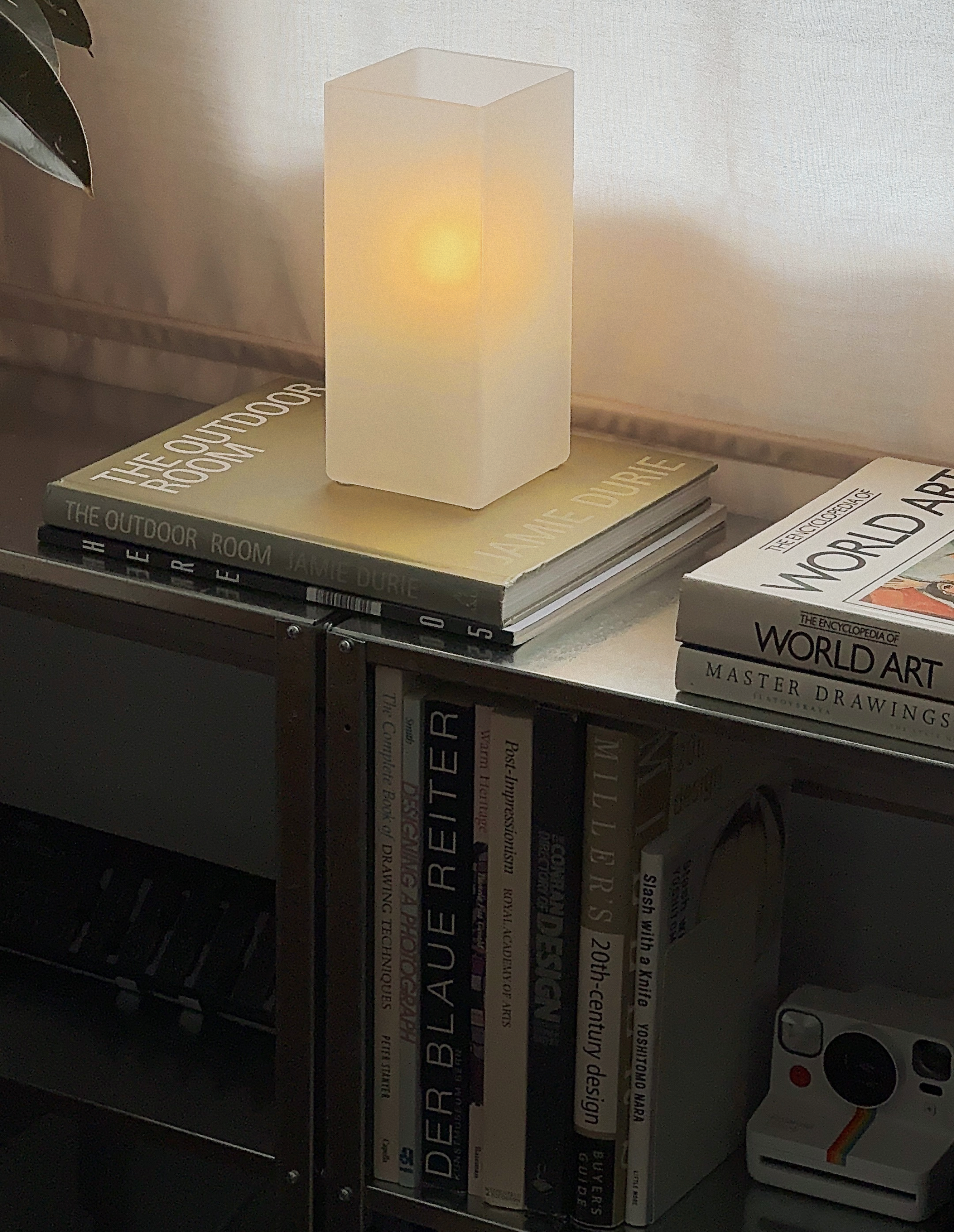 A modern table lamp with a rectangular shade glowing softly, placed on top of a stack of books including 'The Outdoor Room' by Jamie Durie and 'World Art.' Below, a bookshelf contains various art, design, and photography books, with a small camera on the right.