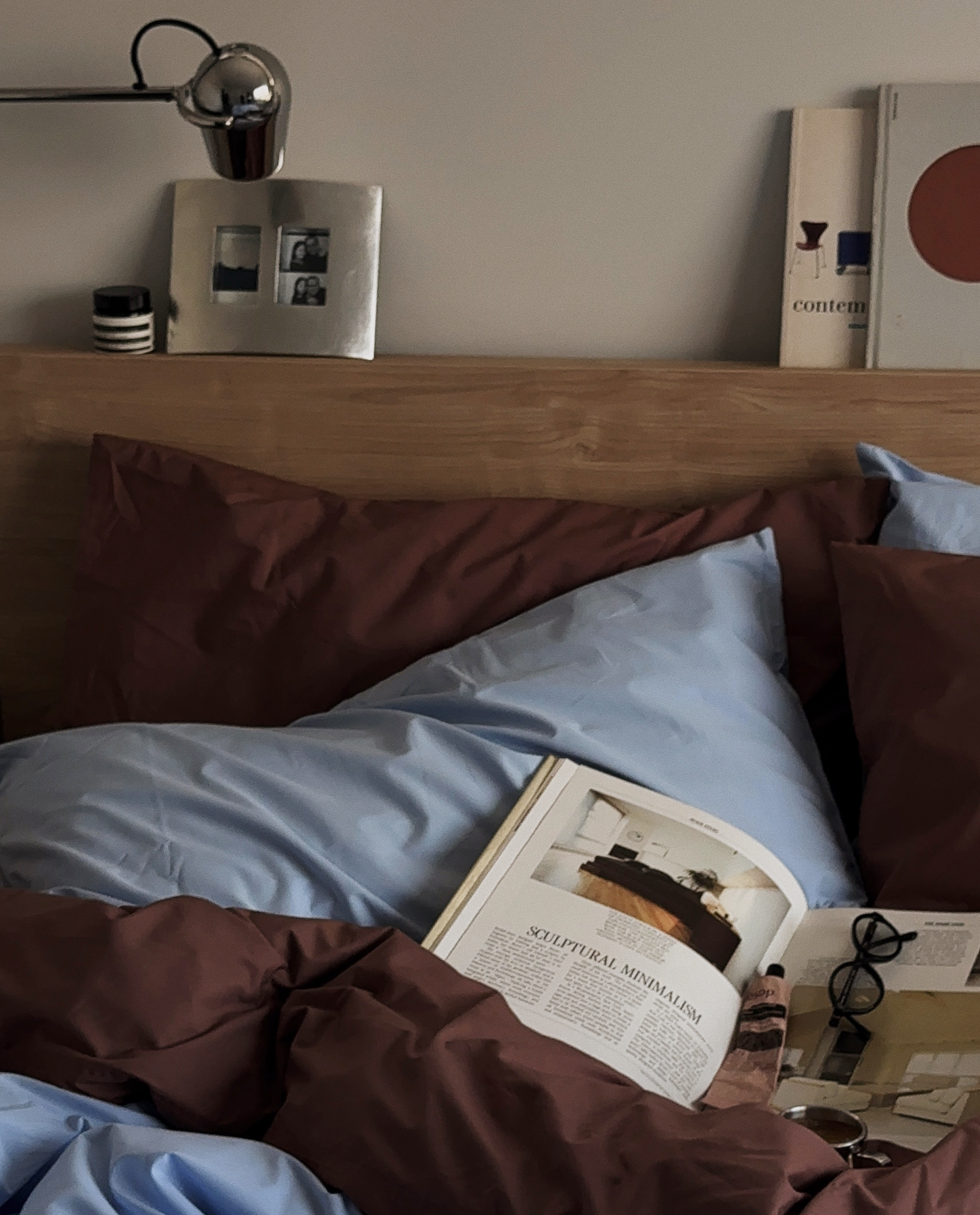 Unmade bed with blue and brown bedding, open magazine, glasses, and a tube of lipstick on the bed, with a wood headboard and wall decorations in the background.