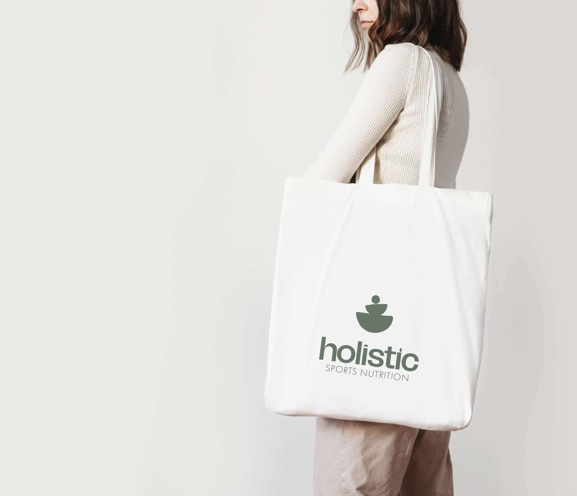 Person holding a large white tote bag with the green logo and text 'holistic SPORTS NUTRITION' on it, standing against a plain background.