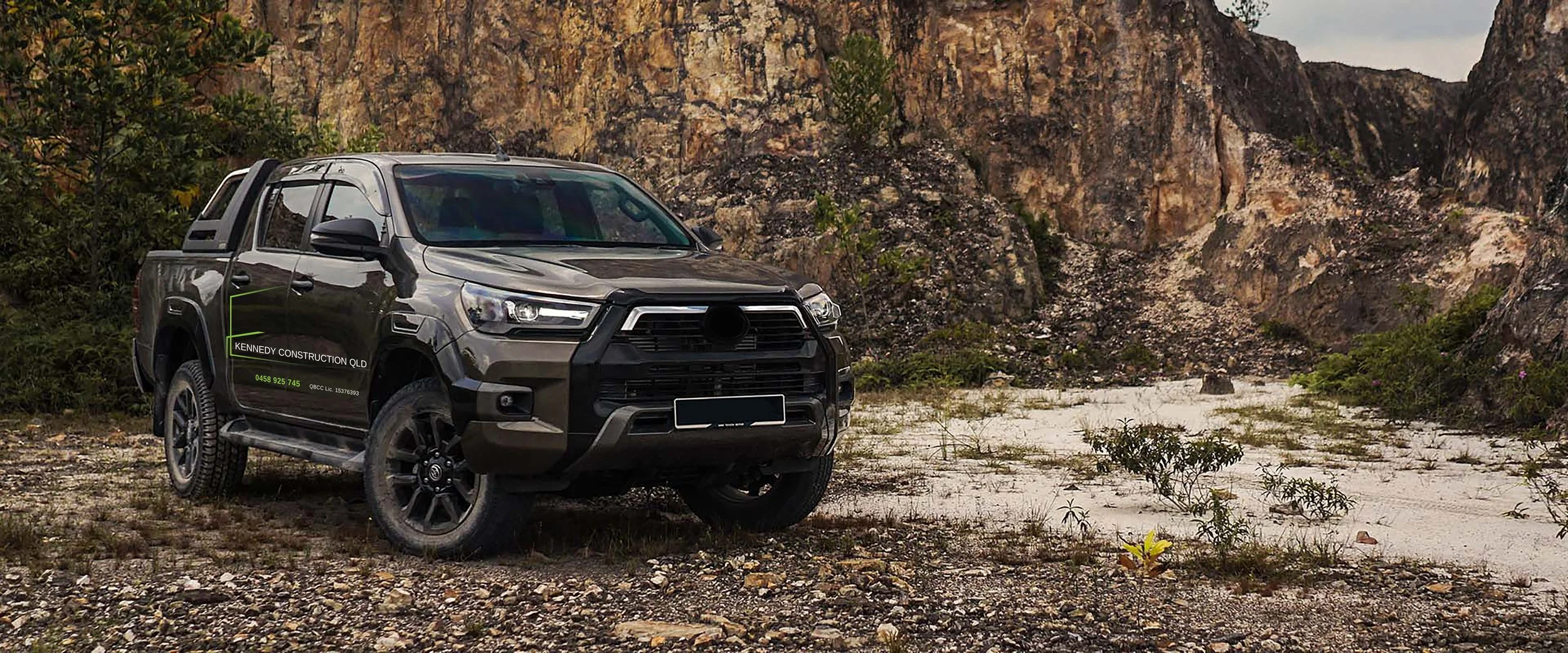 A black pickup truck parked on a rocky, dirt terrain with sparse vegetation, mountains, and a cloudy sky in the background.