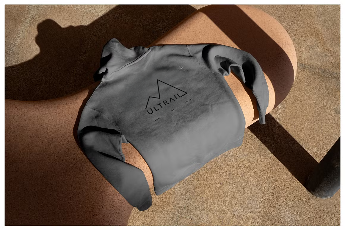 Gray sweatshirt with 'ULTRAIL' logo draped over a person's lap, who is sitting on a bench with sunlight casting shadows on the ground.