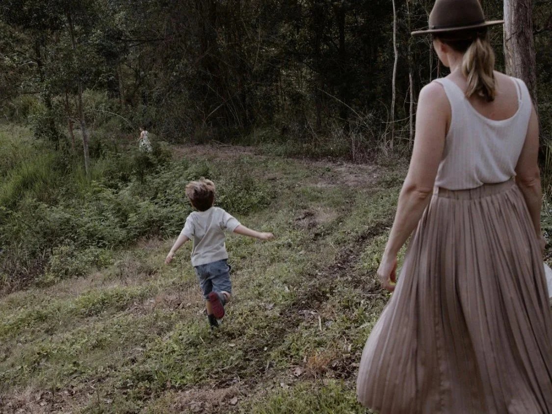 A woman and a young boy running through a wooded area on a dirt trail, with the woman wearing a wide-brimmed hat and a long pleated skirt, and the boy wearing a light-colored shirt and shorts.