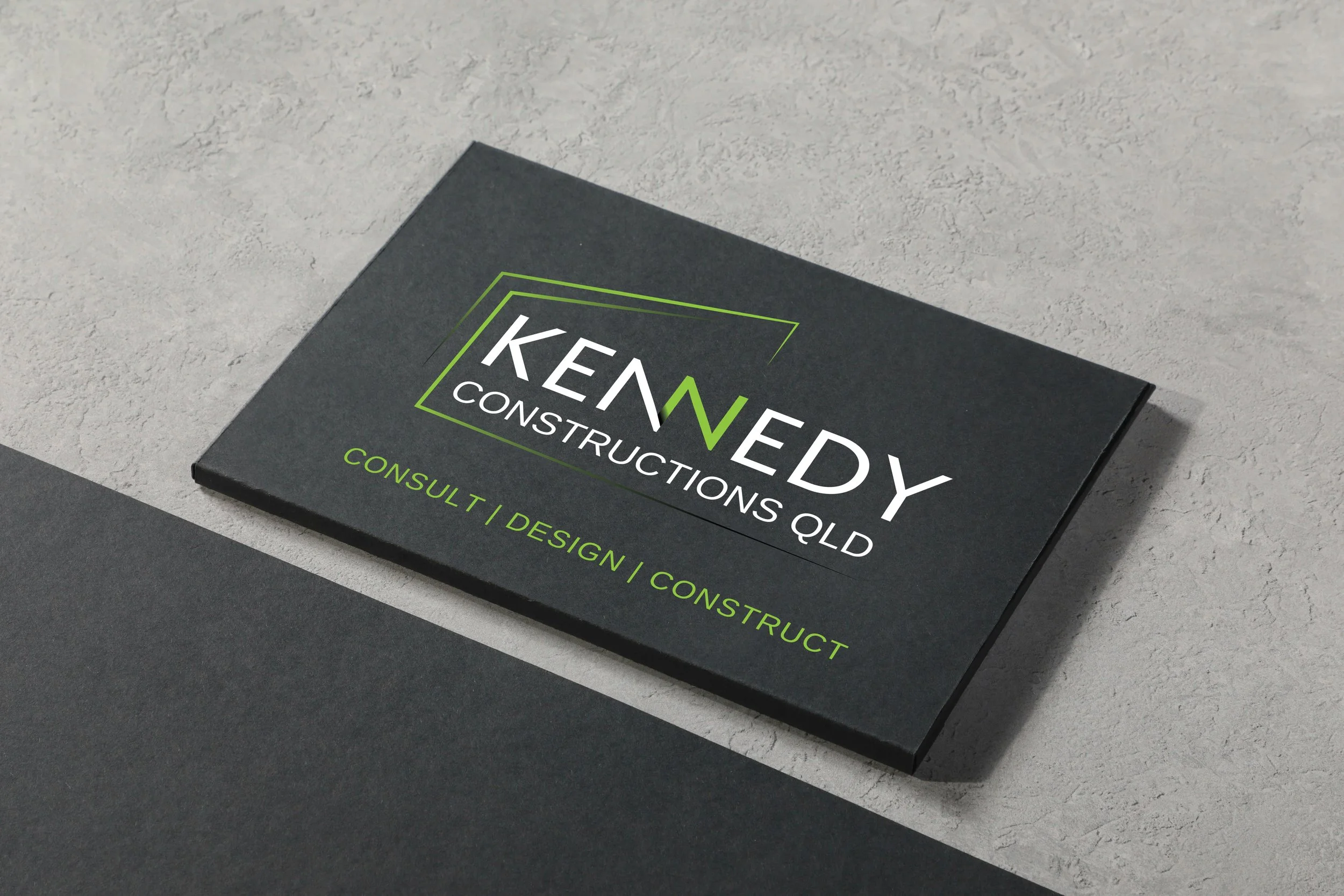 Business card for Kennedy Constructions Queensland with green and white text on a black background, placed on a gray and black surface.