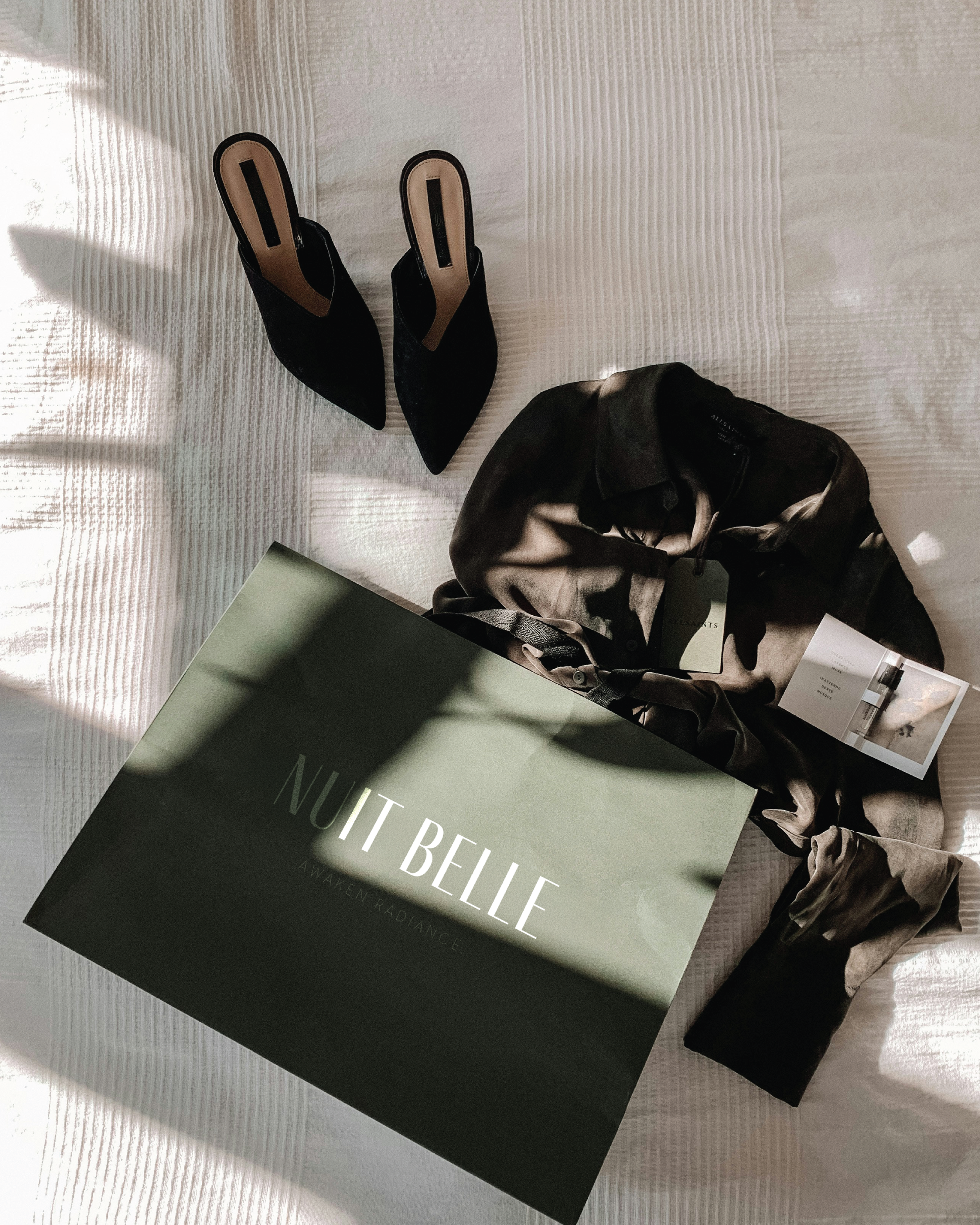 Black high heel shoes, a patterned silk shirt with tags, and a dark green box with "Nuit Belle" written on it, all placed on a white textured surface with sunlight and shadows.