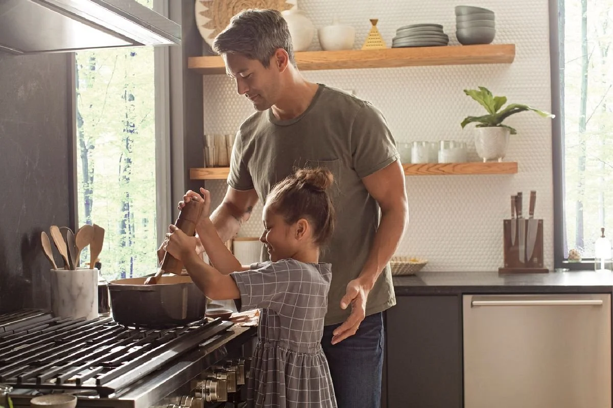 A man and a young girl cooking together in a kitchen, with sunlight streaming through the window.