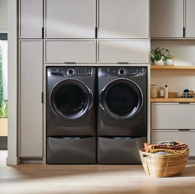 A modern laundry room with a front-loading washer and dryer in dark gray, installed side by side, surrounded by light-colored cabinets and shelves with decorative plants and storage baskets.