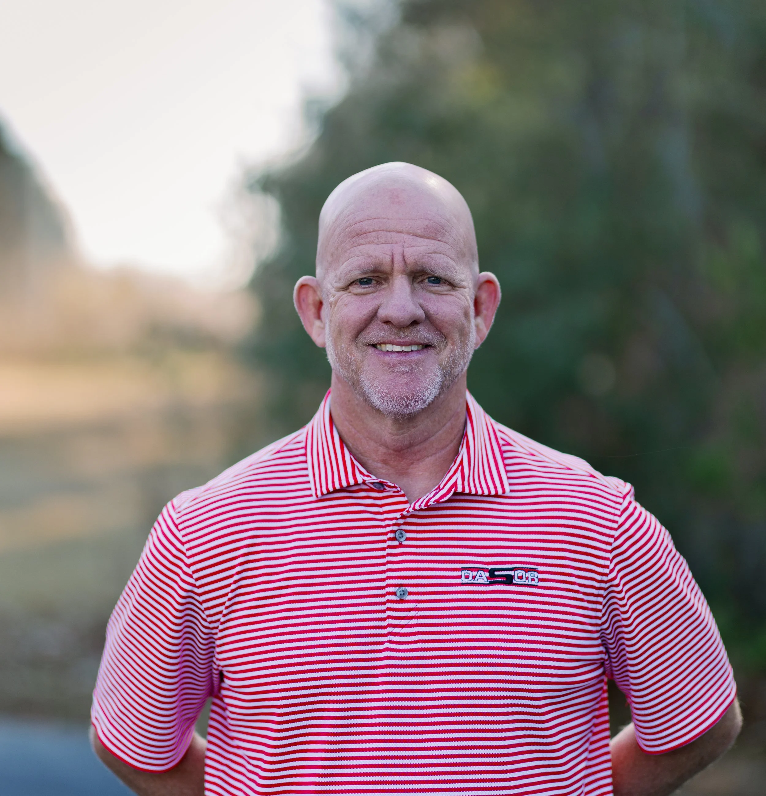 A middle-aged man with a bald head and gray facial hair, wearing a red and white striped polo shirt, standing outdoors with trees and a blurred background.
