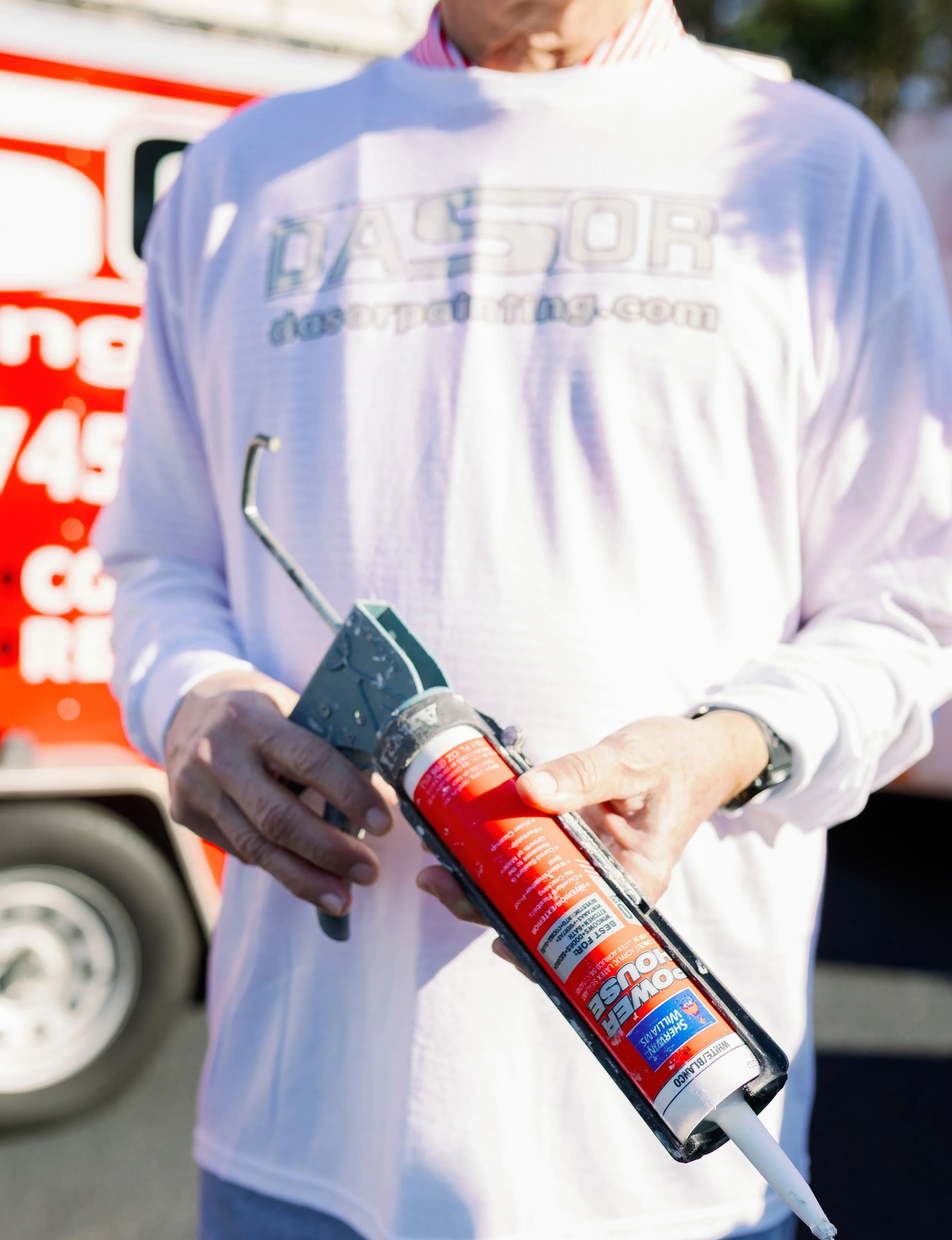 Person holding a tube of silicone sealant with a caulking gun in an outdoor setting.