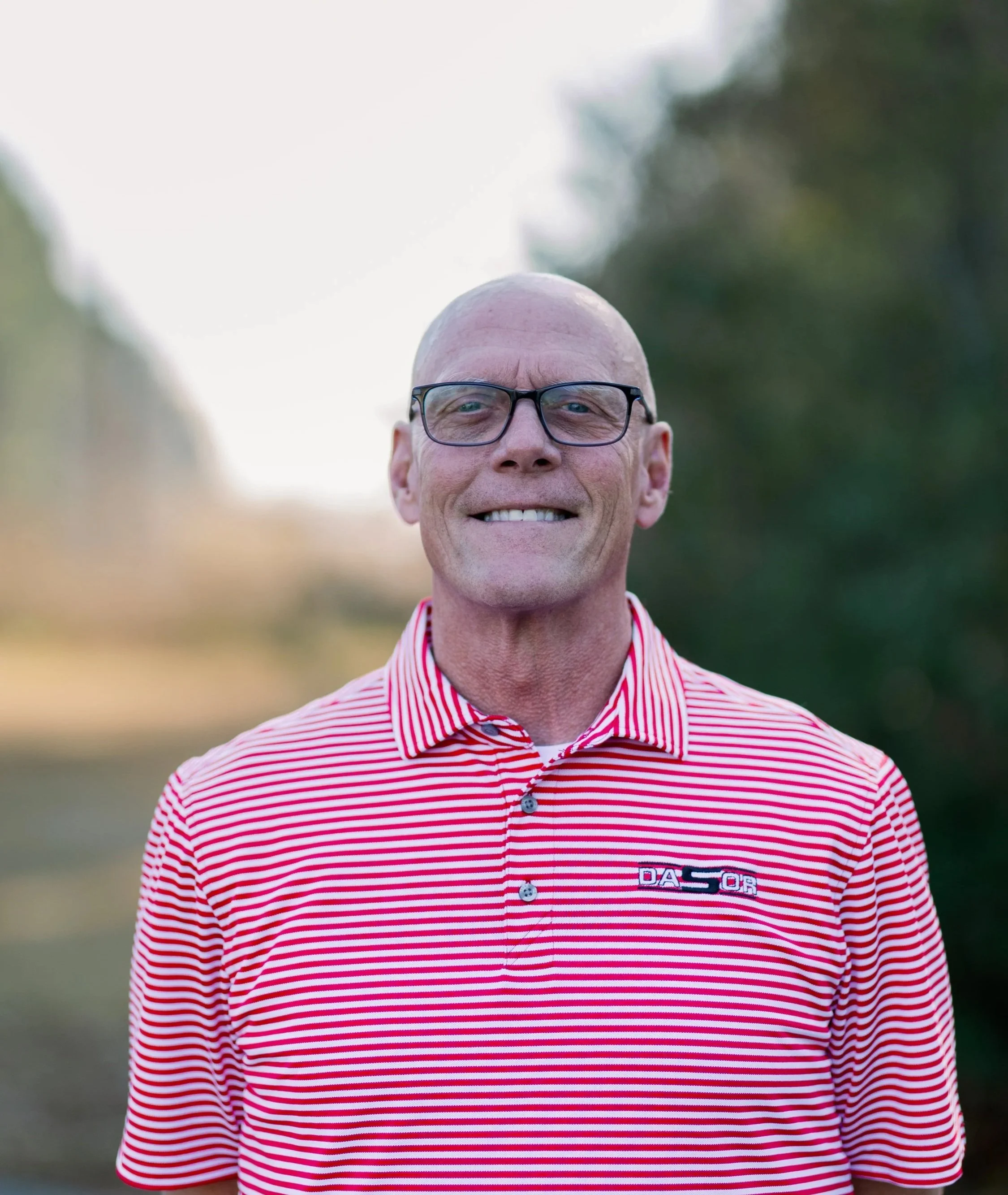 Portrait of a young man with short hair, glasses, smiling, wearing a black long-sleeved shirt, standing against a plain white background.
