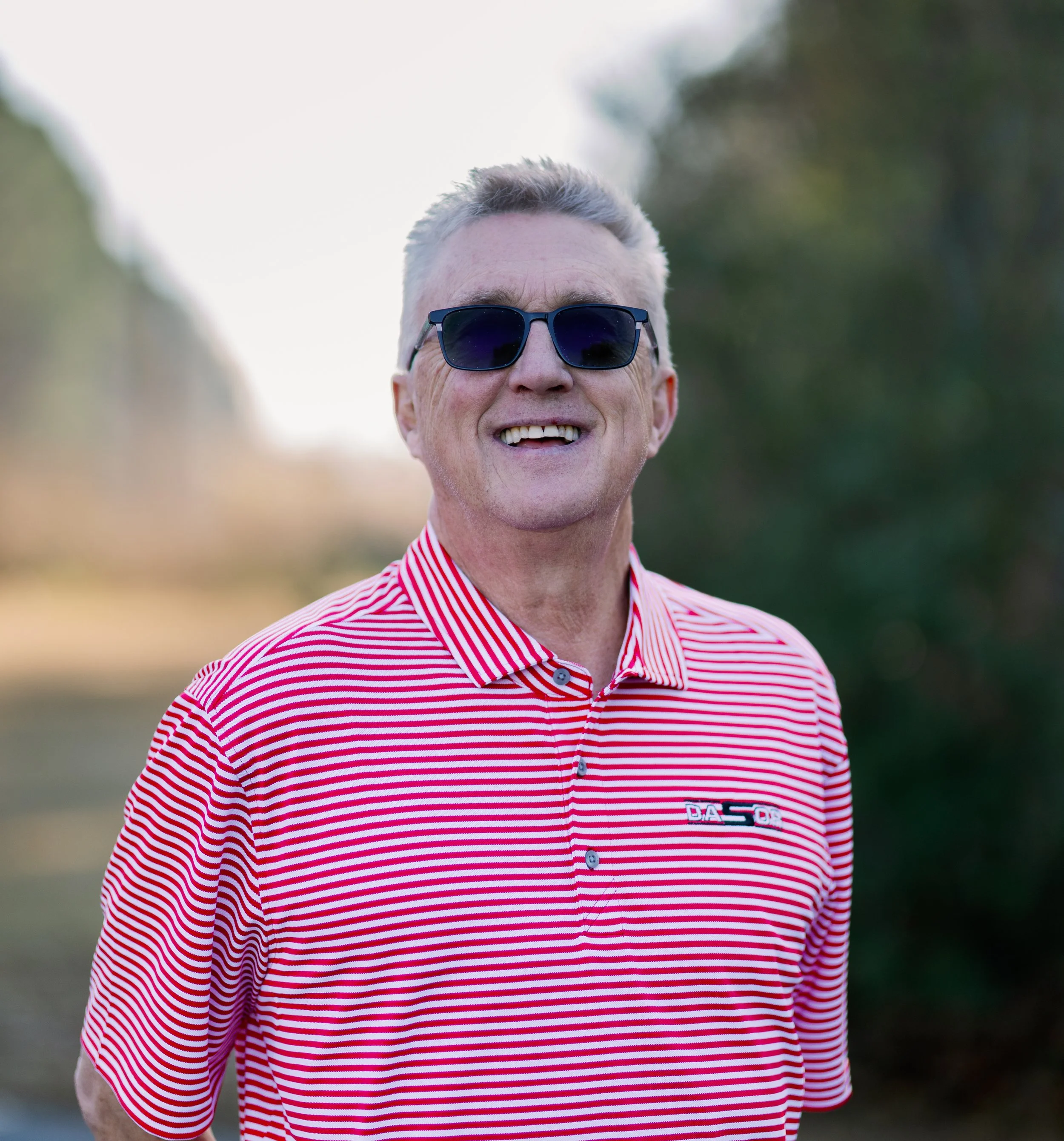 A middle-aged man with gray beard and short gray hair, wearing a brown polo shirt and white pants, sitting on a wooden bench against a plain wall.