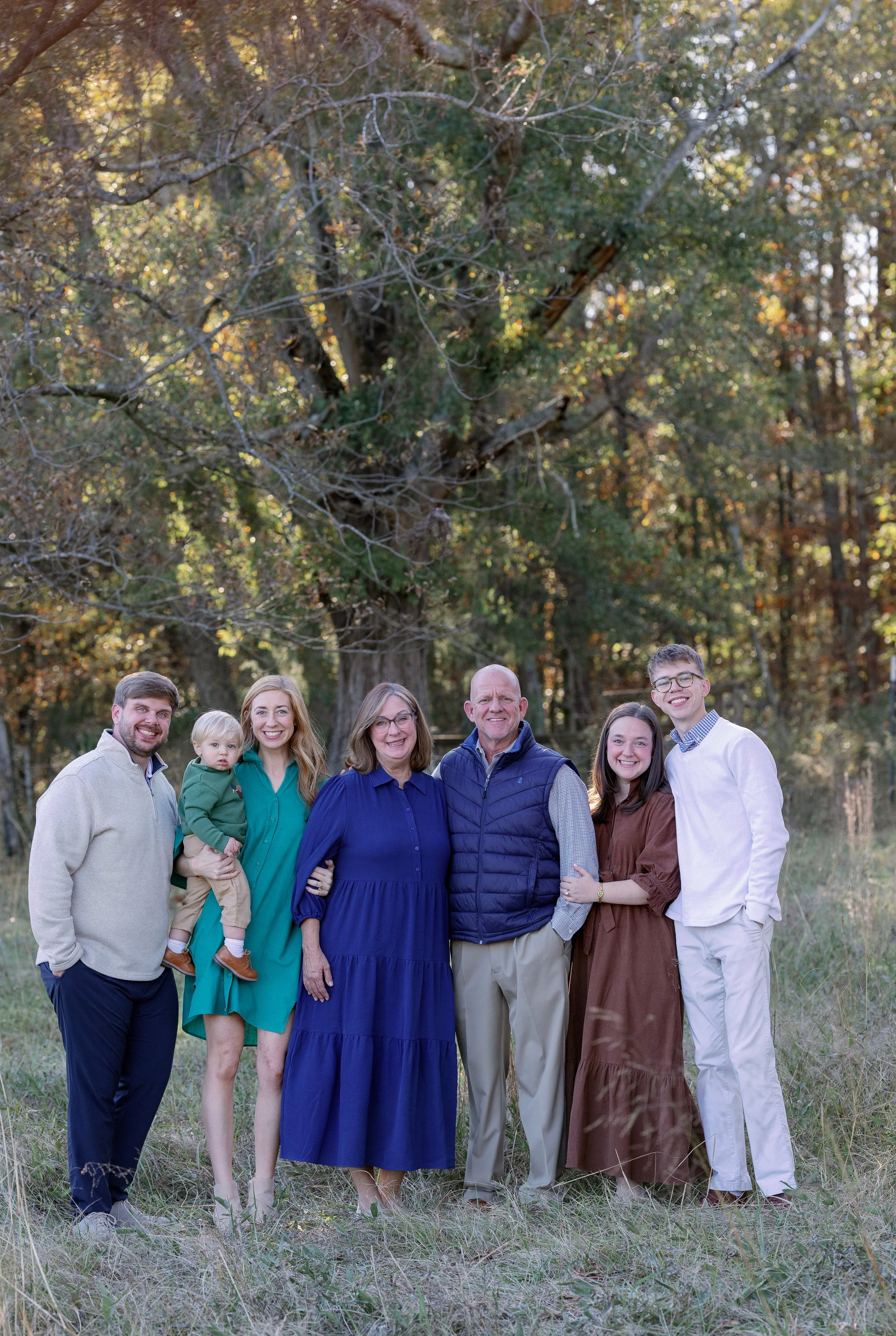 Family of seven standing outdoors in a wooded area during fall, smiling at the camera. The group includes a man, woman, an elderly woman, an elderly man, and three younger individuals, one holding a small child.