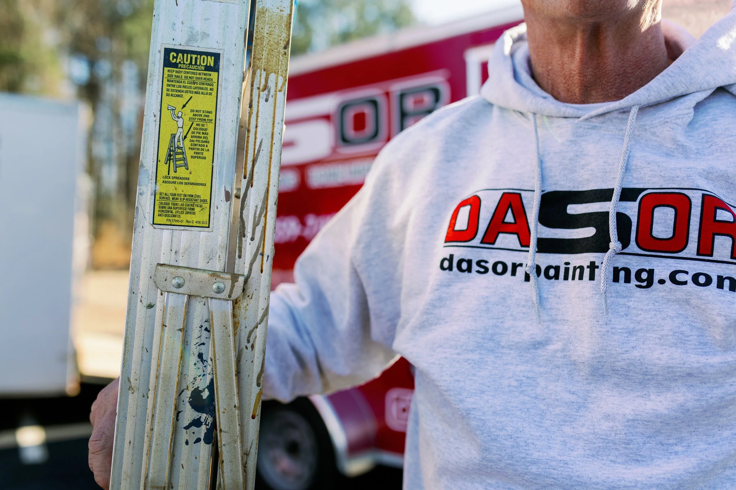Person wearing a gray hoodie with a logo and text, holding a step ladder with a caution sticker on it. Background includes a red vehicle with partially visible white text.