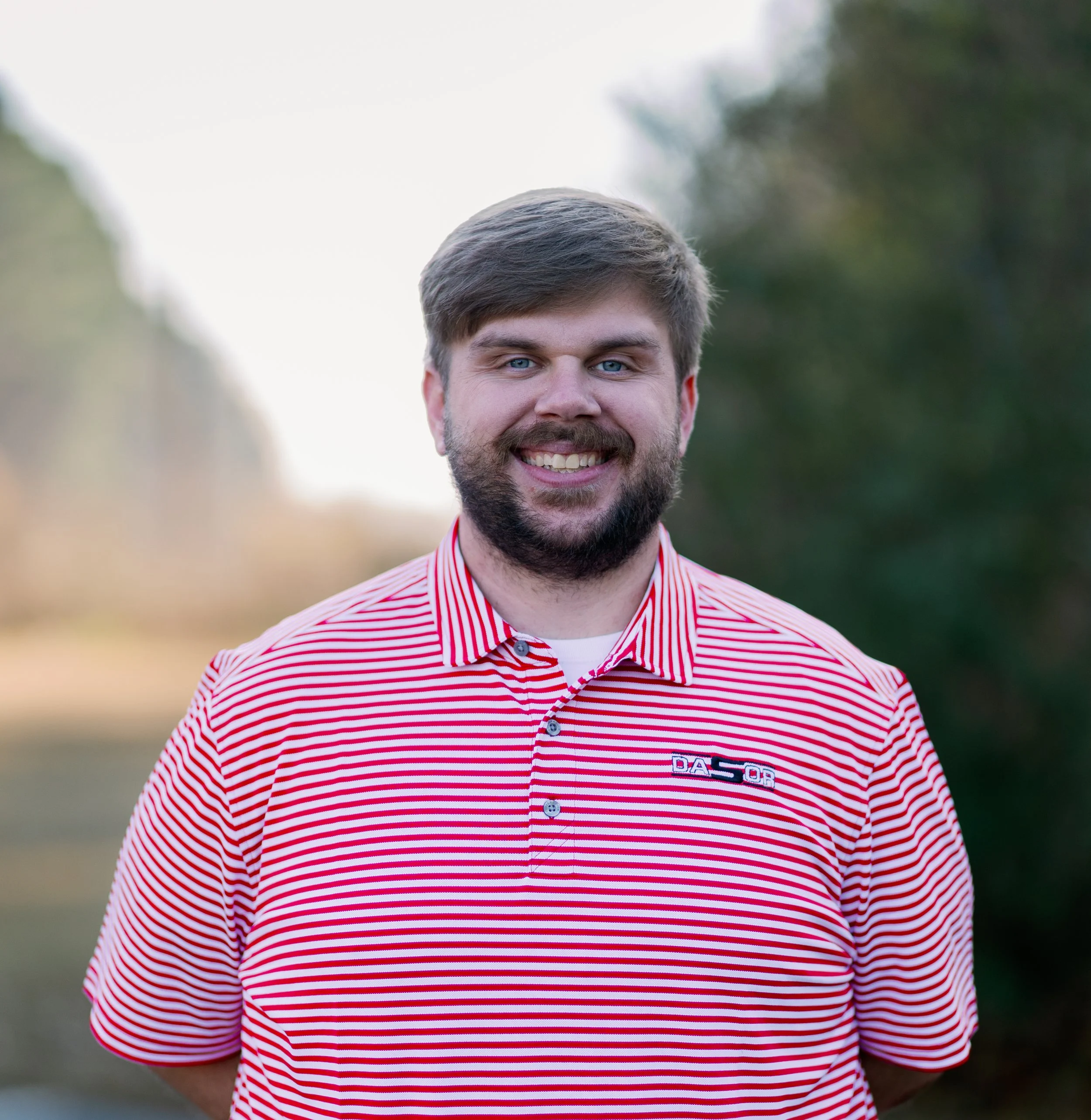 A smiling man with a beard and short hair standing outdoors in a forested area, wearing a red and white striped polo shirt.
