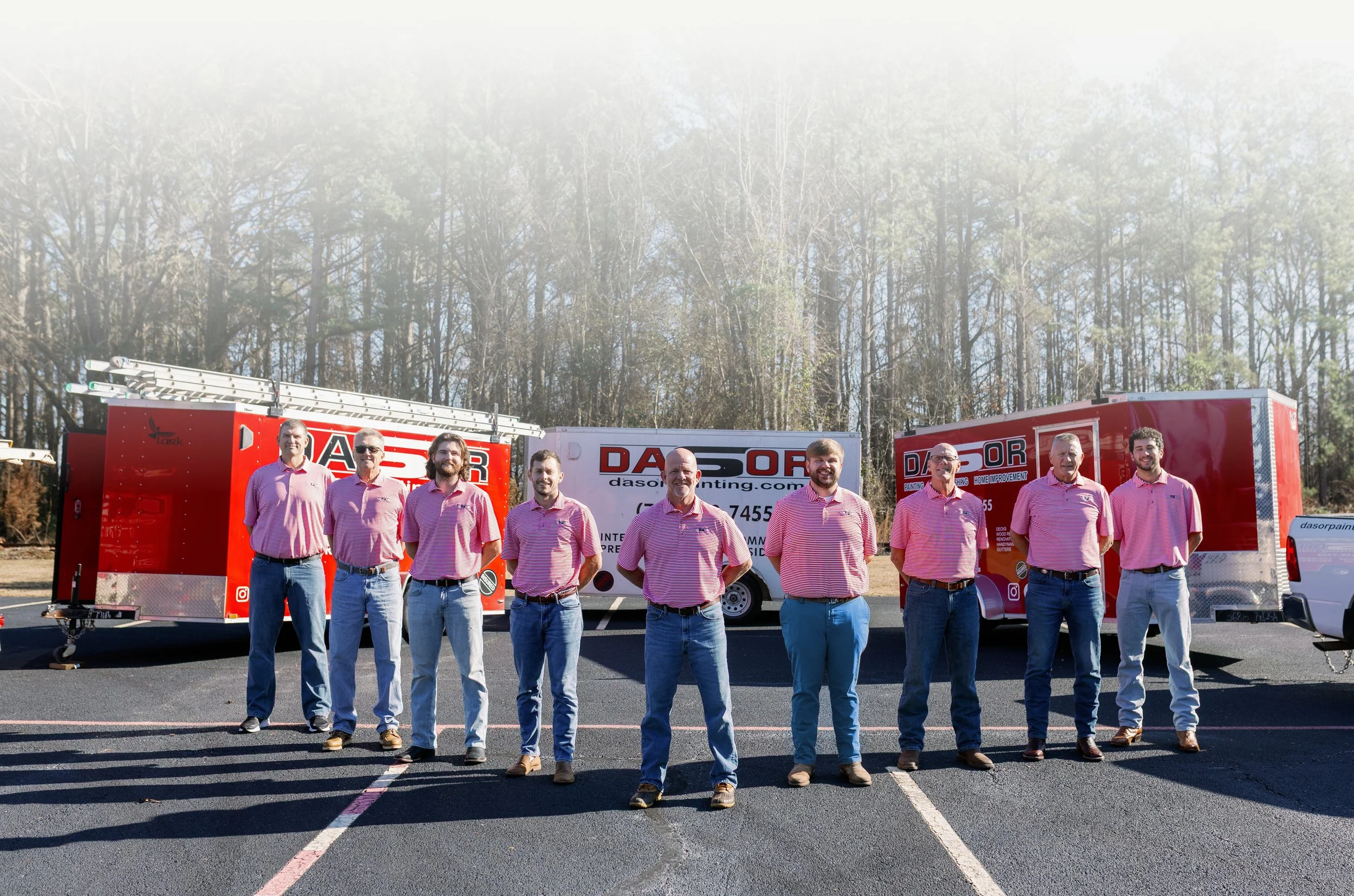 Group of nine men standing in front of red and white Dasor painting service trucks, outdoors on a paved parking lot with trees in the background.