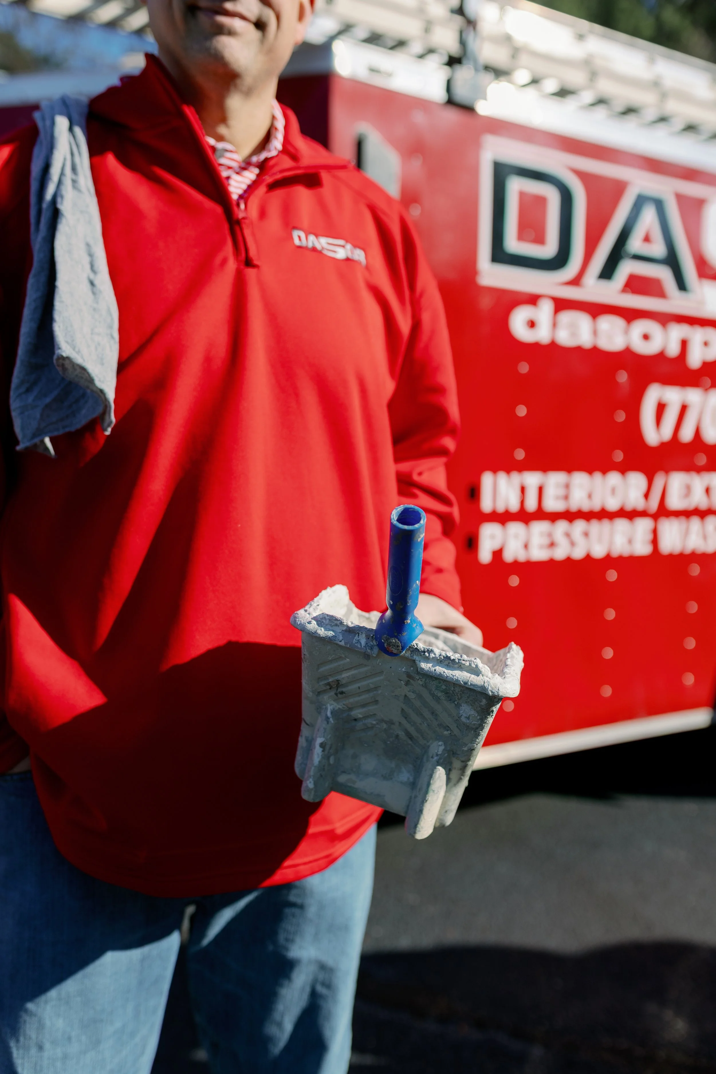 A man wearing a red jacket with a towel over his shoulder holding a paint roller covered in white paint in front of a red truck with business details.