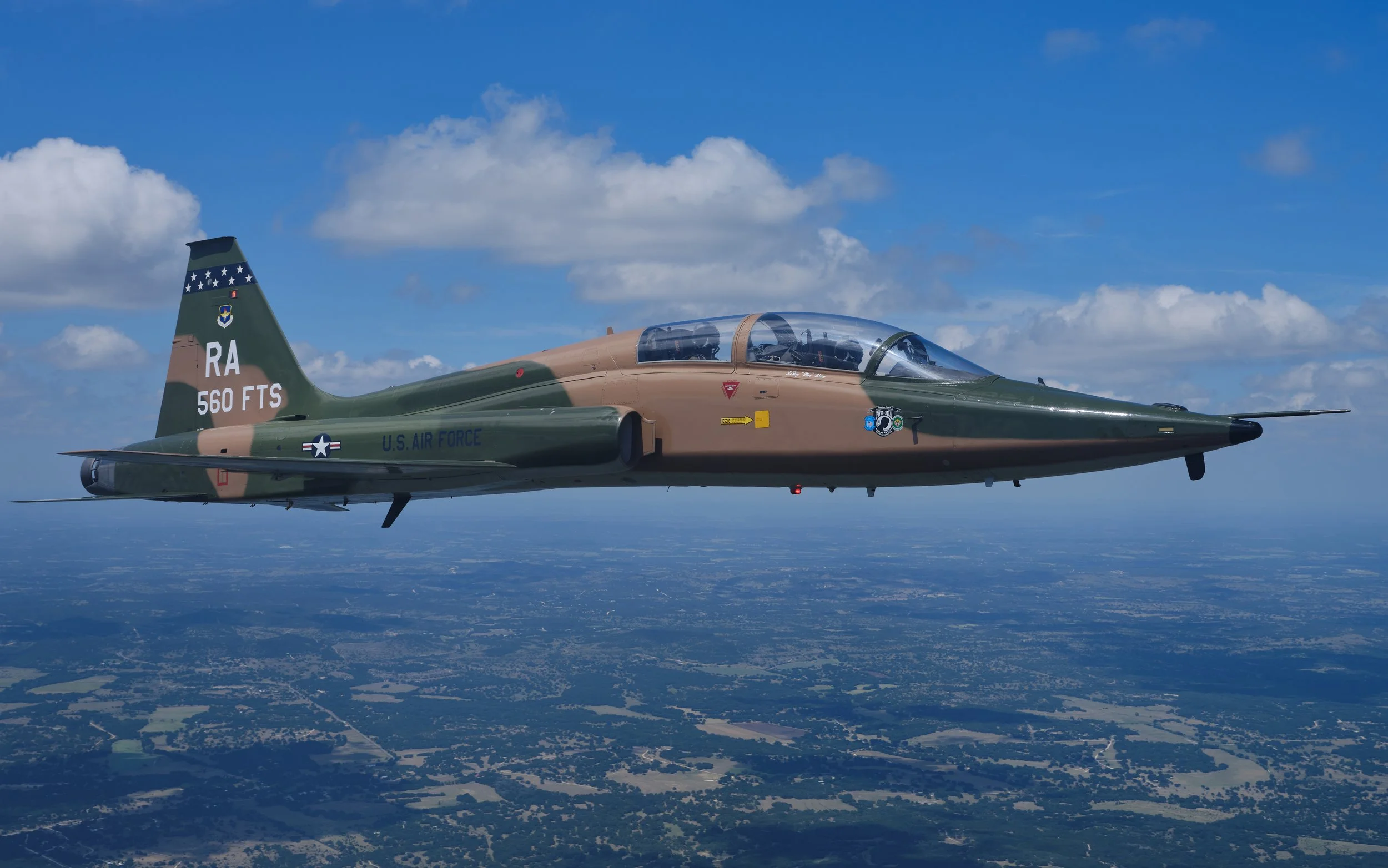 A military aircraft flying high above a landscape with blue sky and scattered clouds.