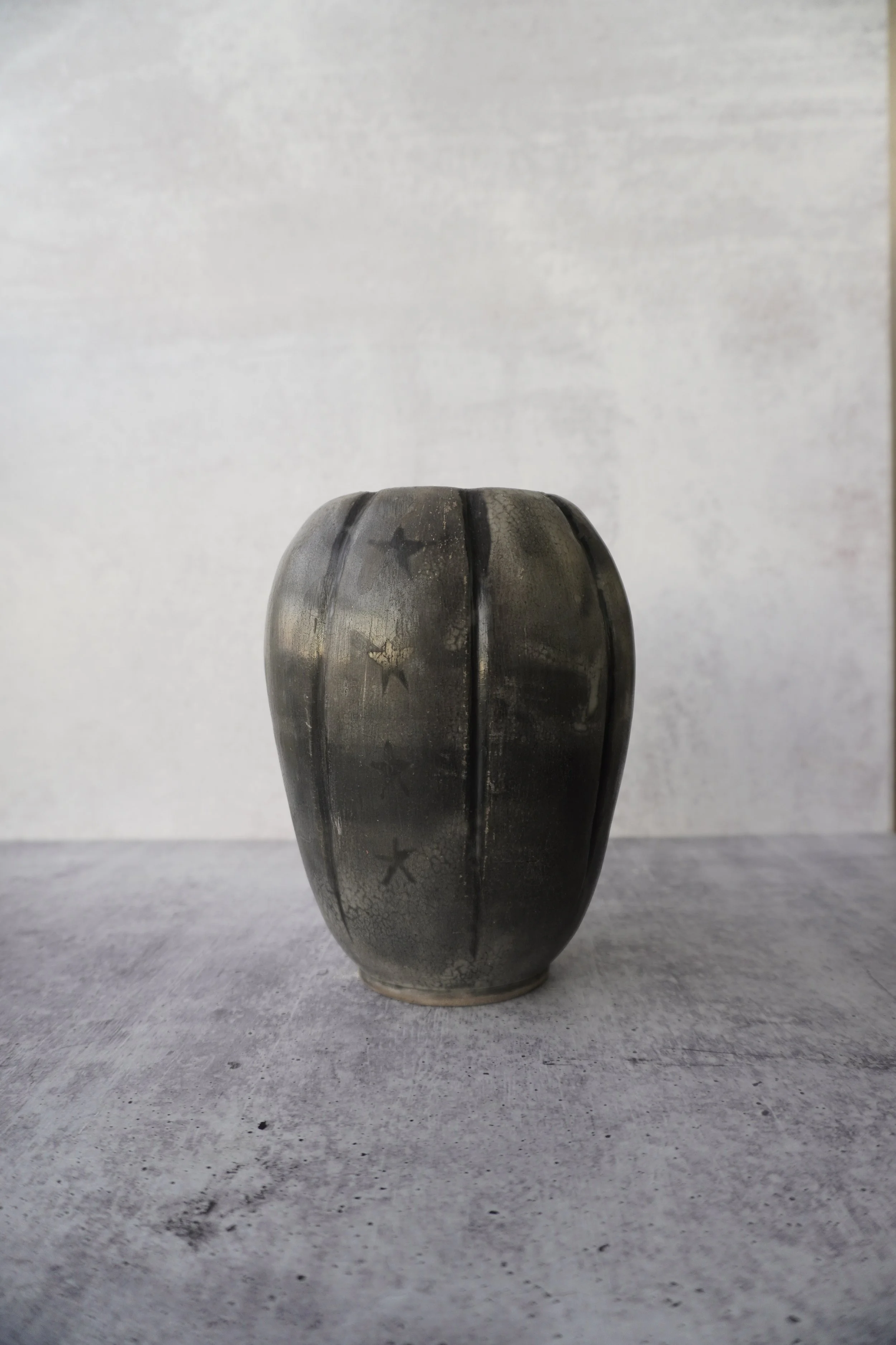 A dark, metallic vase with star patterns, standing on a gray textured surface against a plain light background.