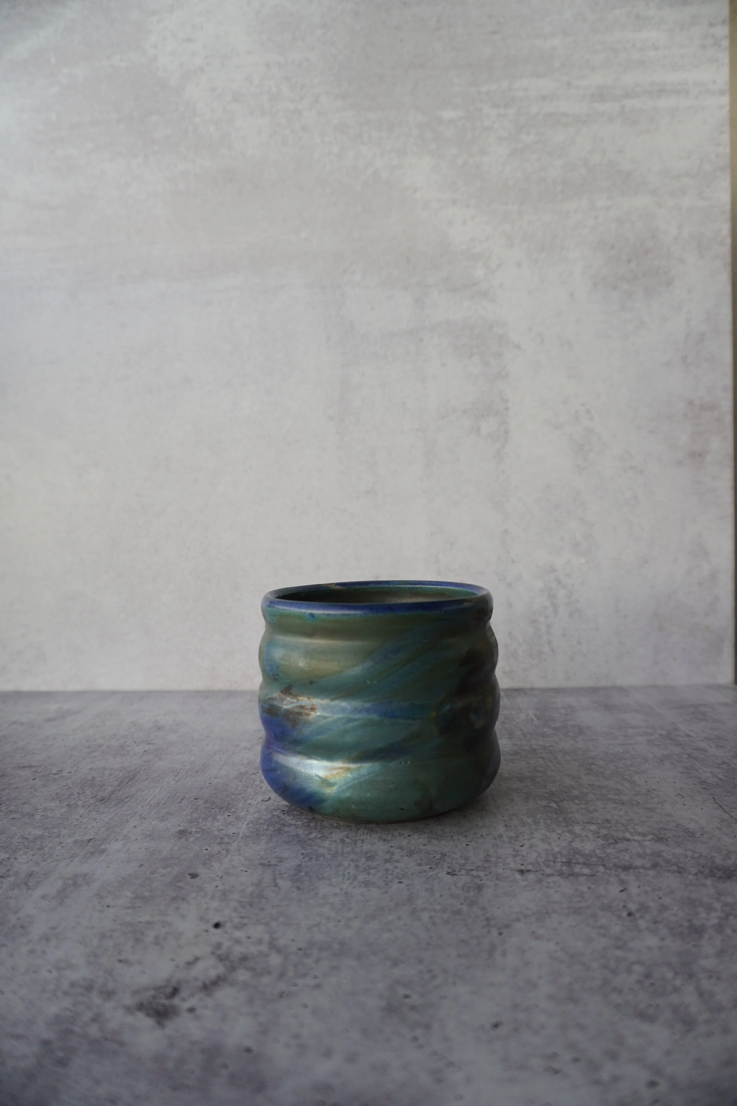 A small, swirl-patterned ceramic pot with blue, green, and beige colors on a gray surface against a gray wall background.