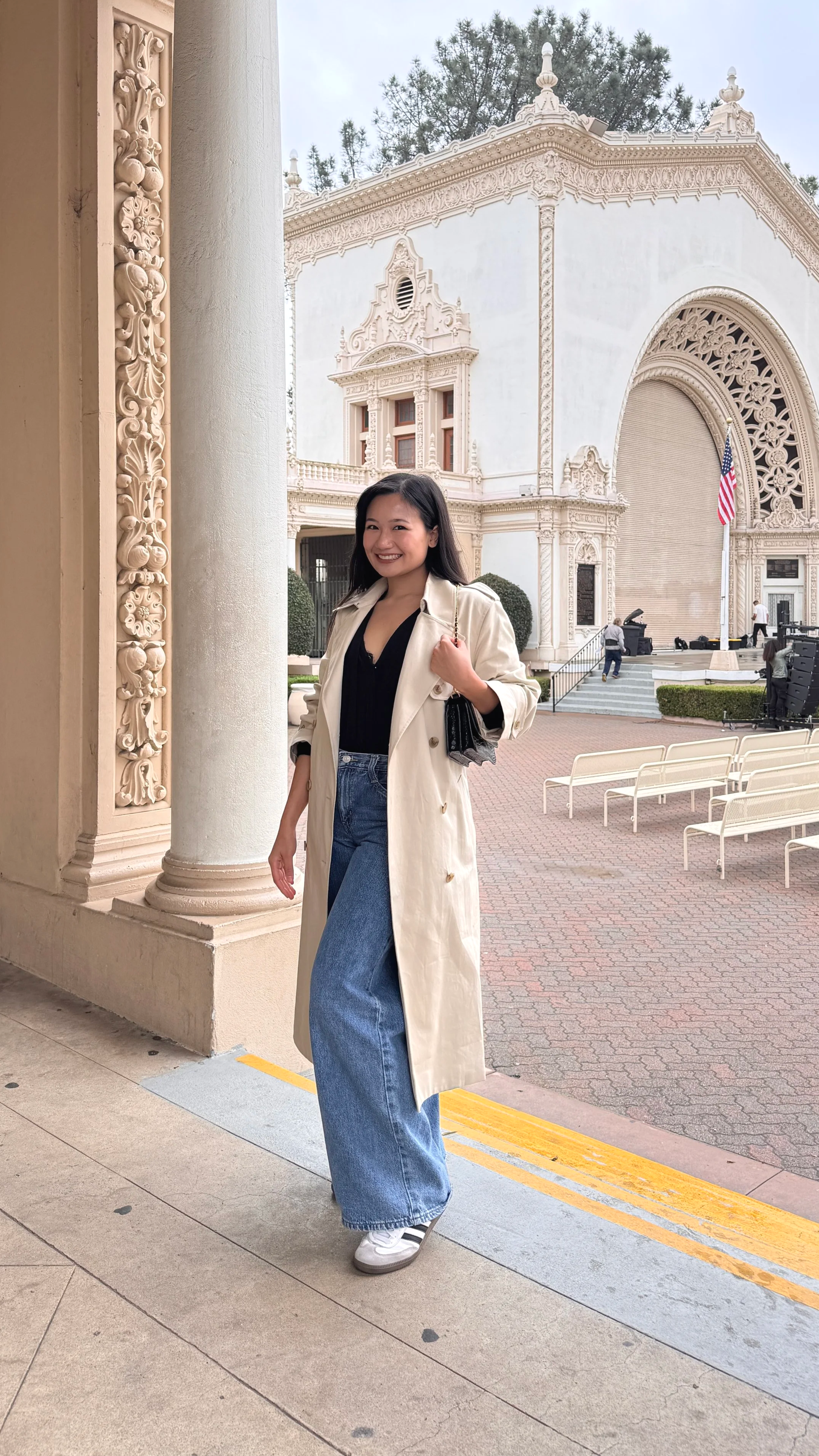 A woman standing outside a historic building with ornate architecture, wearing a beige trench coat, black top, and wide-leg jeans, smiling at the camera.