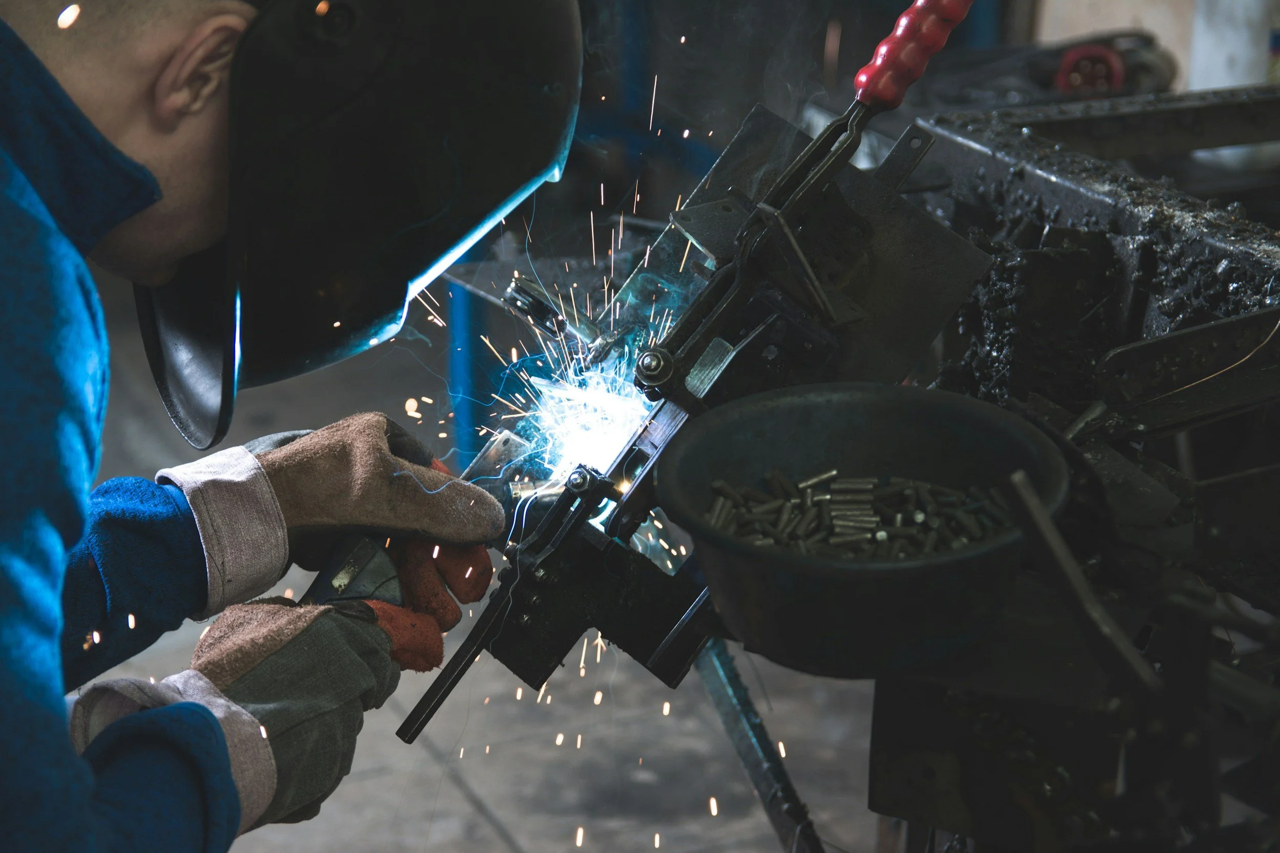 A person wearing protective gloves and helmet welding metal parts in a workshop, with sparks flying from the welding process.