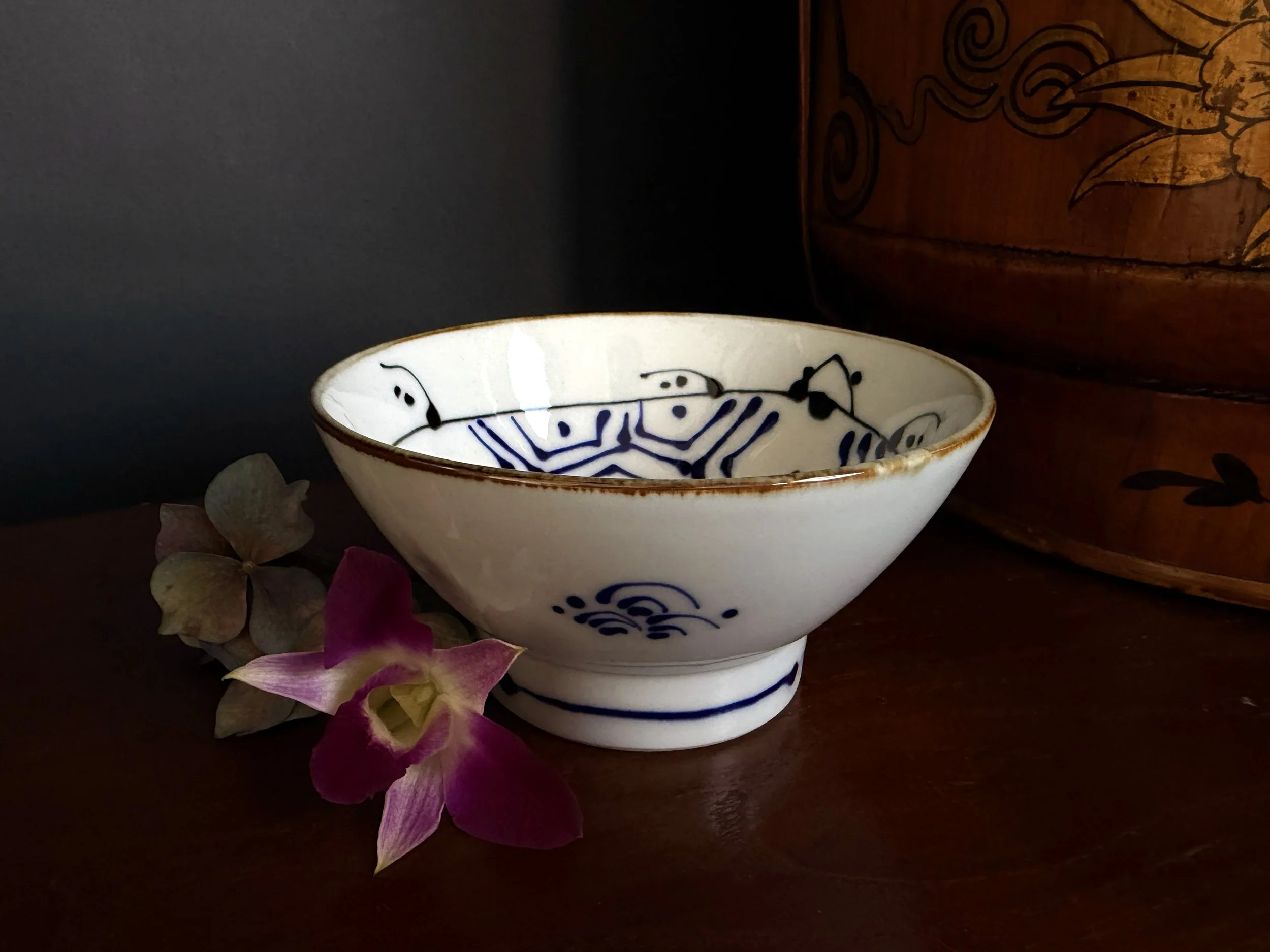 A white ceramic bowl with blue patterns, placed on a dark surface with a pink orchid flower nearby, and a wooden box in the background.