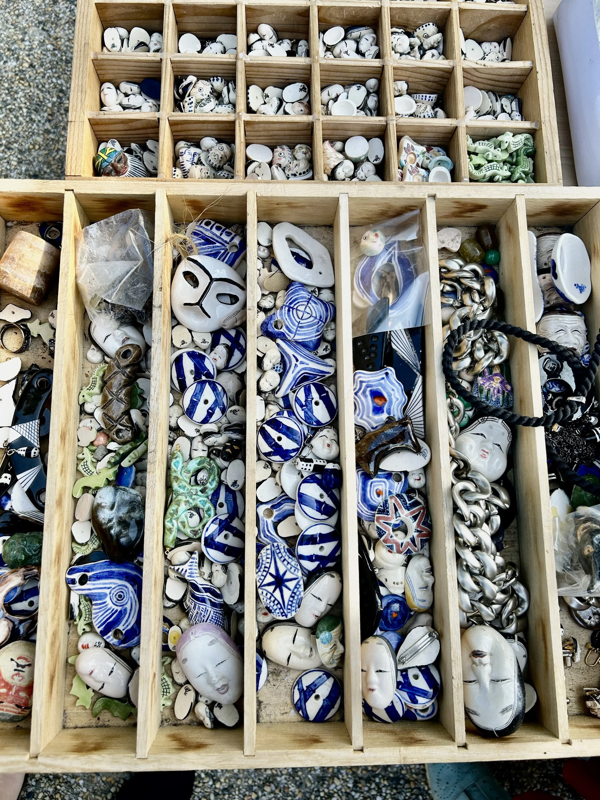 A wooden display case with multiple compartments holding small decorative items such as painted ceramic faces, beads, and jewelry.