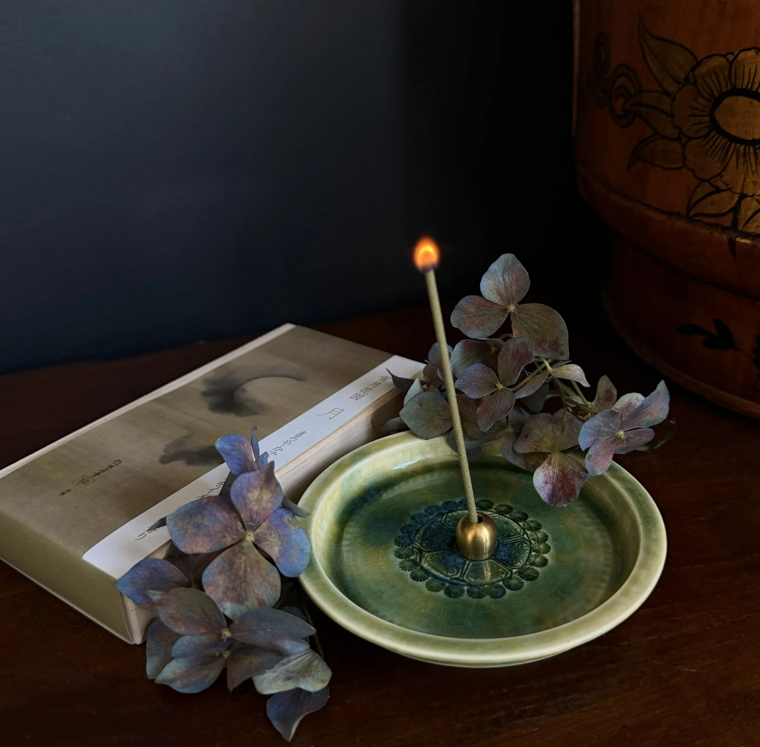 A lit matchstick in a small green and blue ceramic dish with dried hydrangea flowers, a book with a black and white cover, and a wooden container with floral and bird designs, all on a dark wooden surface.