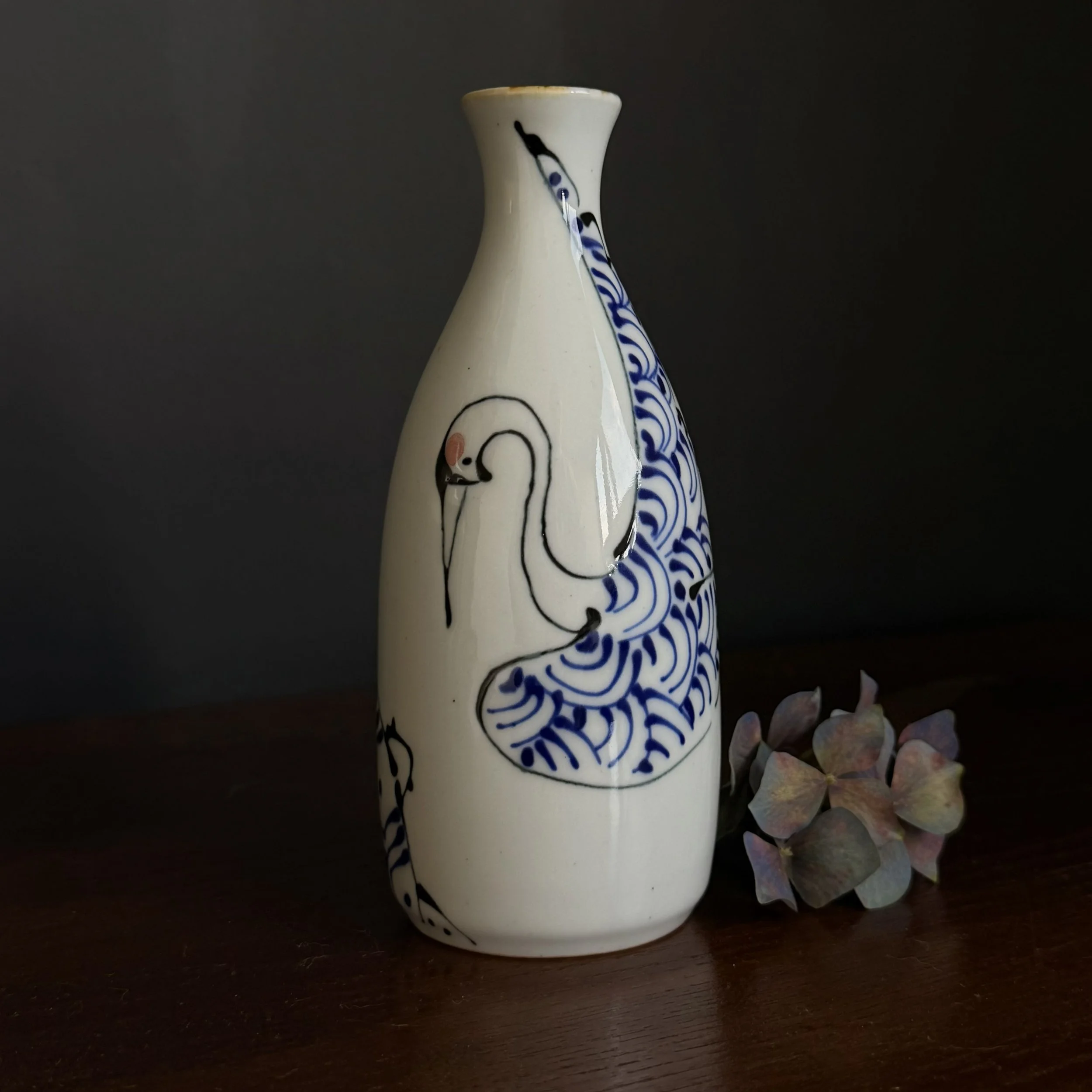 A ceramic vase with a hand-painted black and blue design of a crane and water waves, placed on a wooden surface with dried flowers beside it, against a dark background.
