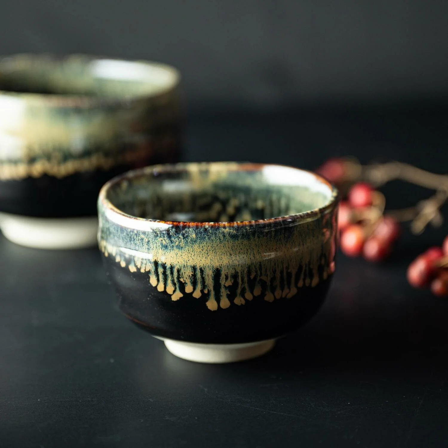 Close-up of a glazed ceramic matcha bowl with a dark exterior and multicolored drip glaze pattern, set on a black surface with a blurred bowl in the background and red berries to the right.
