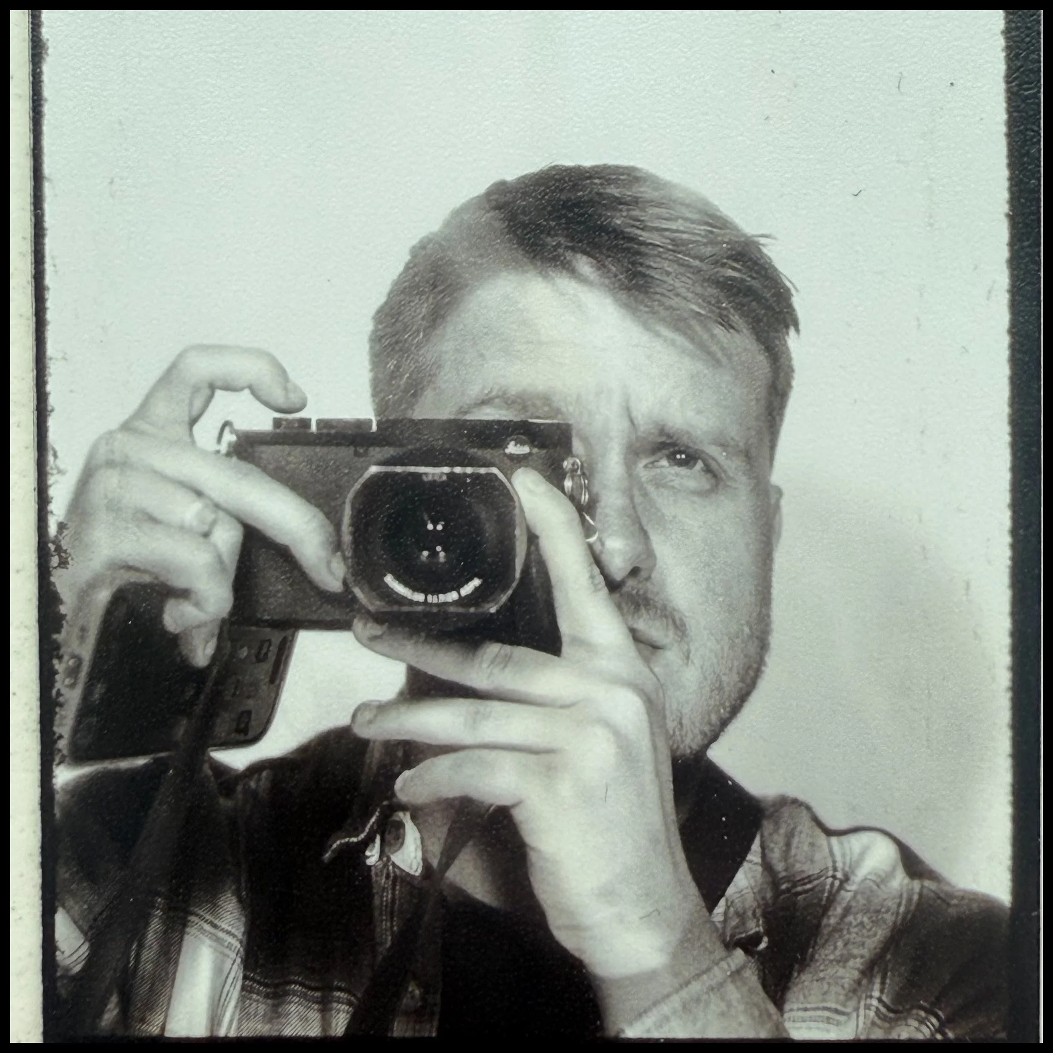 Black and white photo of a man taking a selfie in a mirror, holding a camera with one hand, with glasses and a plaid shirt.