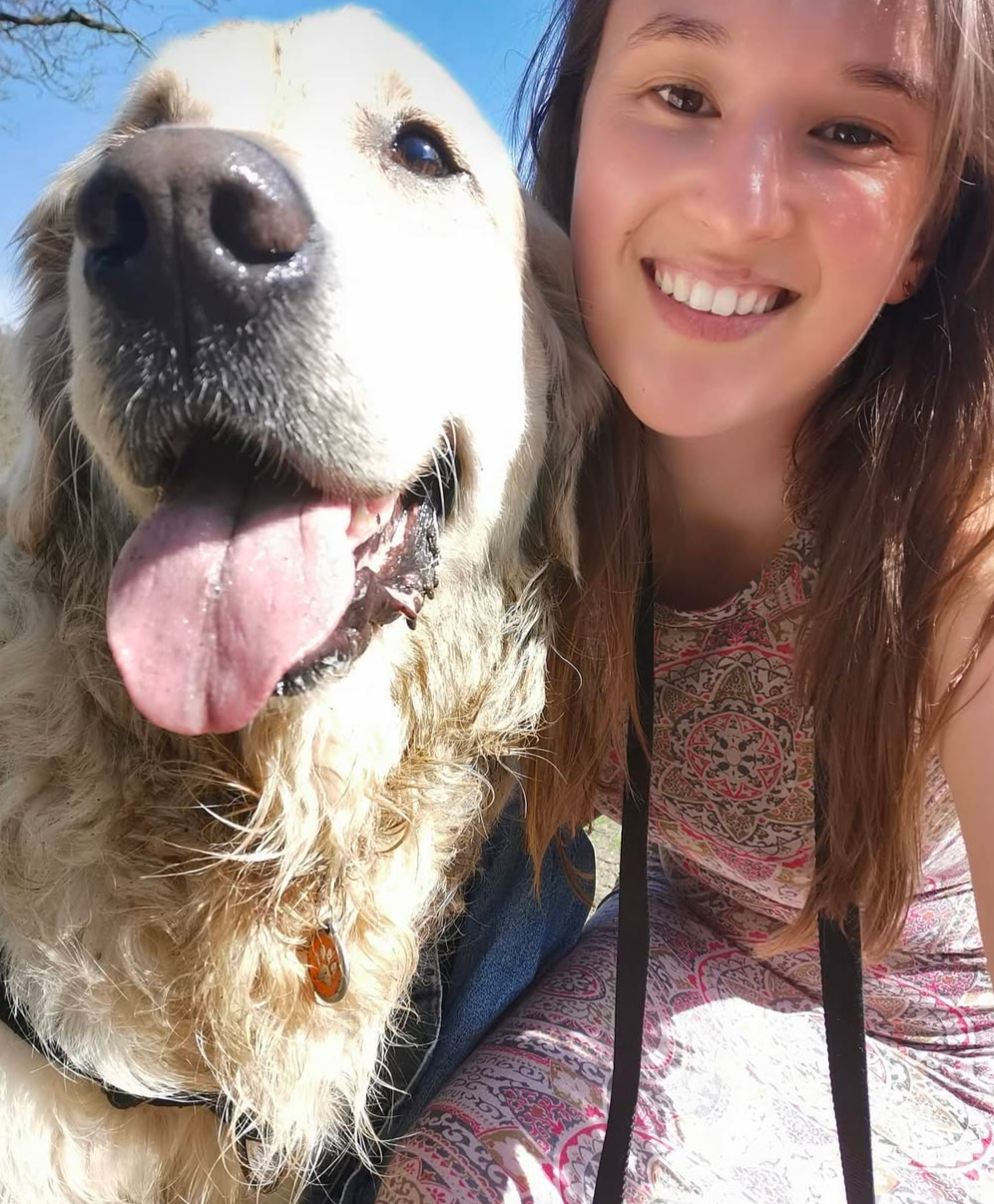 A woman smiling beside a large, fluffy dog with its tongue out, outdoors on a sunny day.