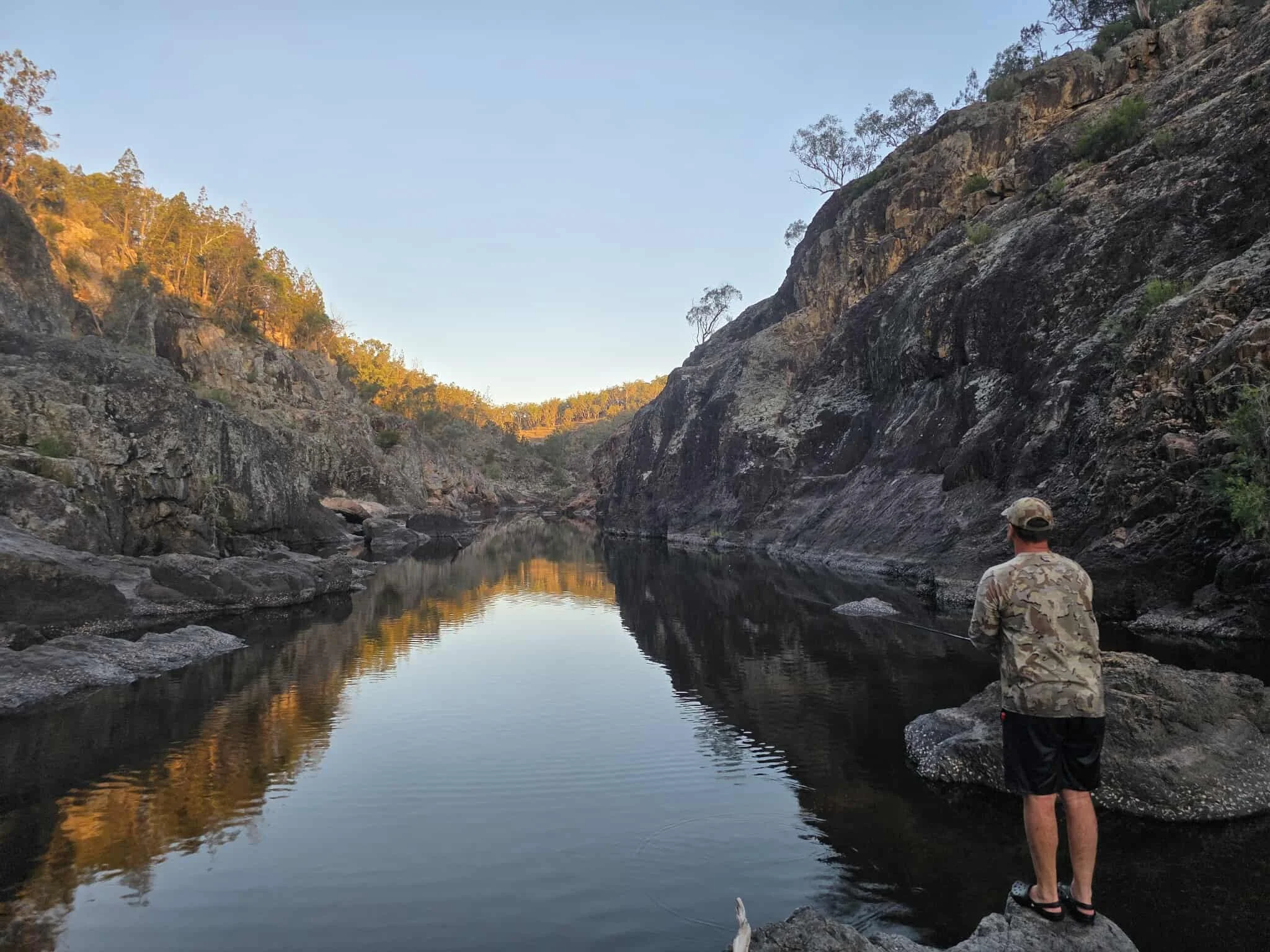 A person fishing on the rocks beside a calm river in a canyon, with rocky cliffs and trees in the background.