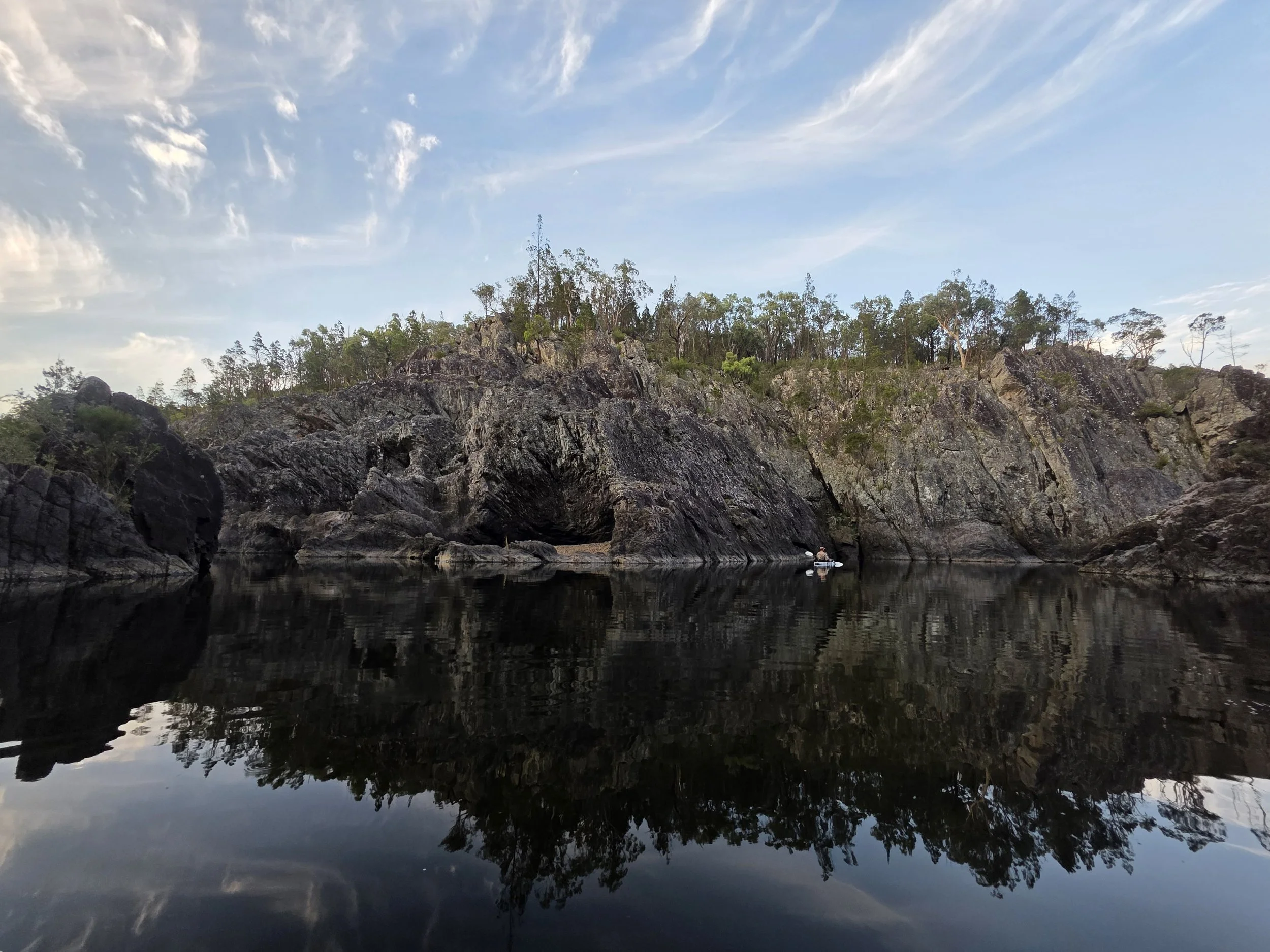 A calm body of water reflecting rocky cliffs and green trees under a partly cloudy sky.