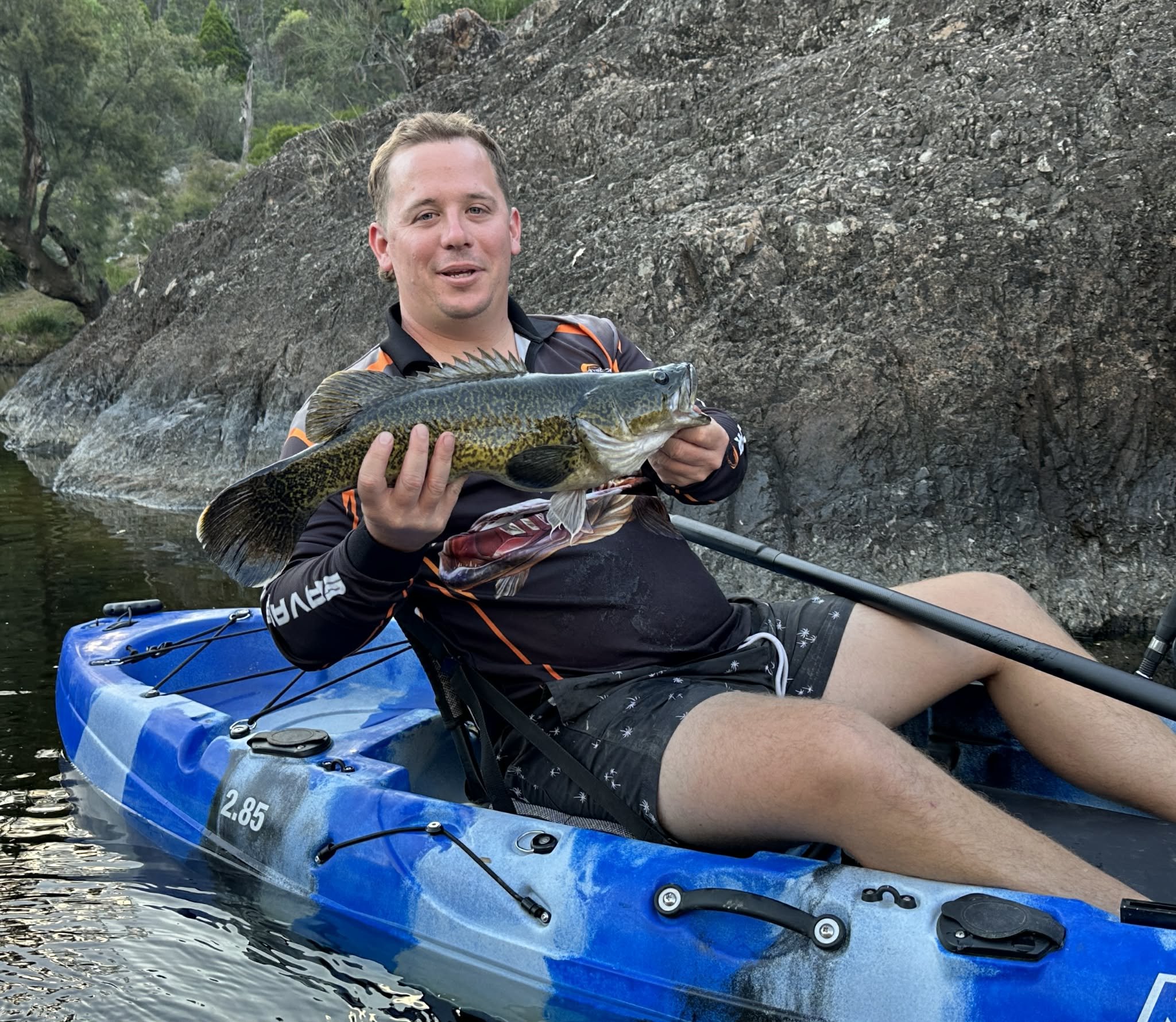 A man sitting in a blue kayak holding a large fish, with a rocky shoreline and trees in the background.