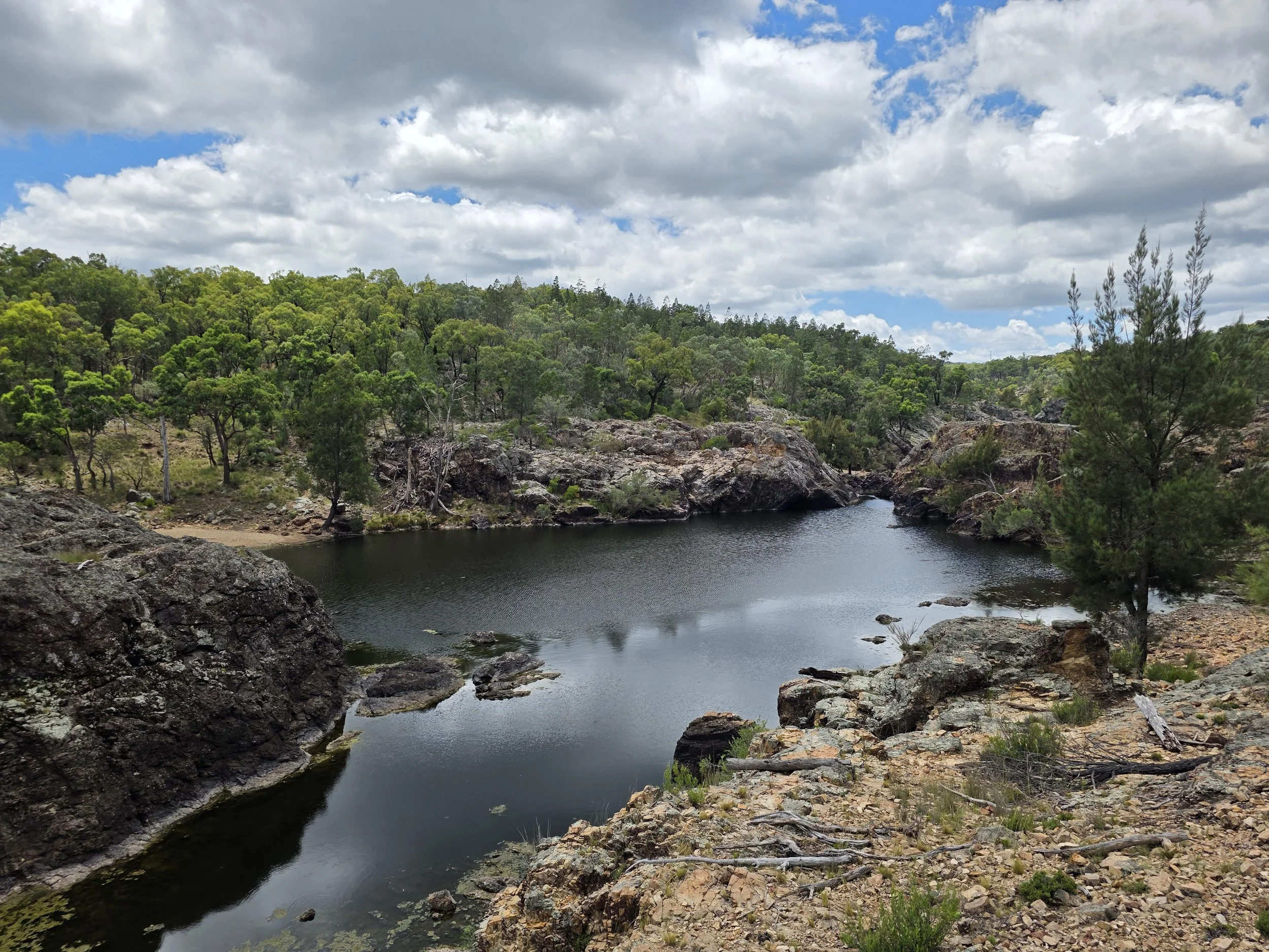 A landscape of a small river or pond surrounded by rocky terrain and green trees under a cloudy sky.