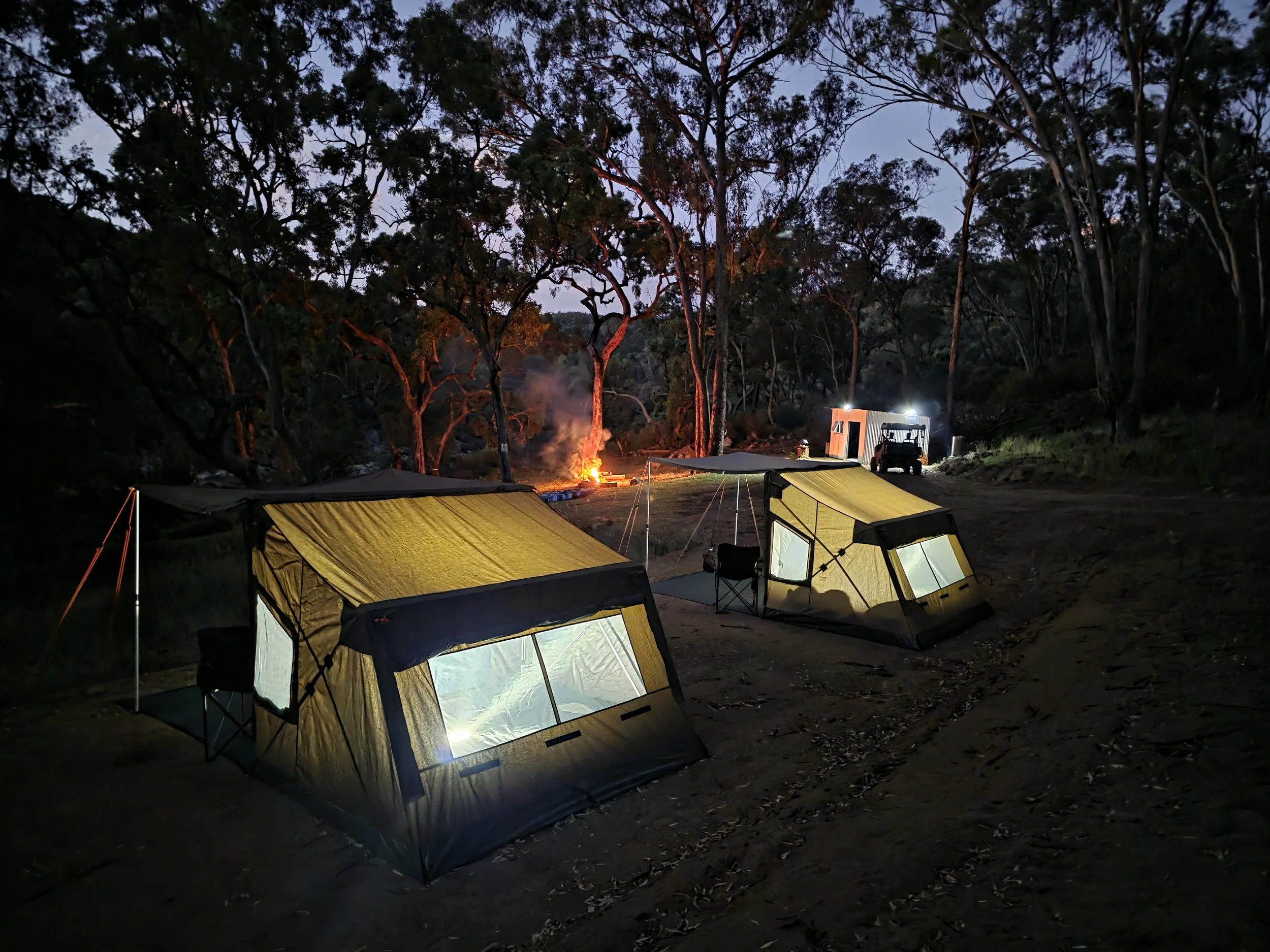 Nighttime scene of a campsite in a forest with two illuminated tents, a small fire burning in the background, and trees surrounding the area.